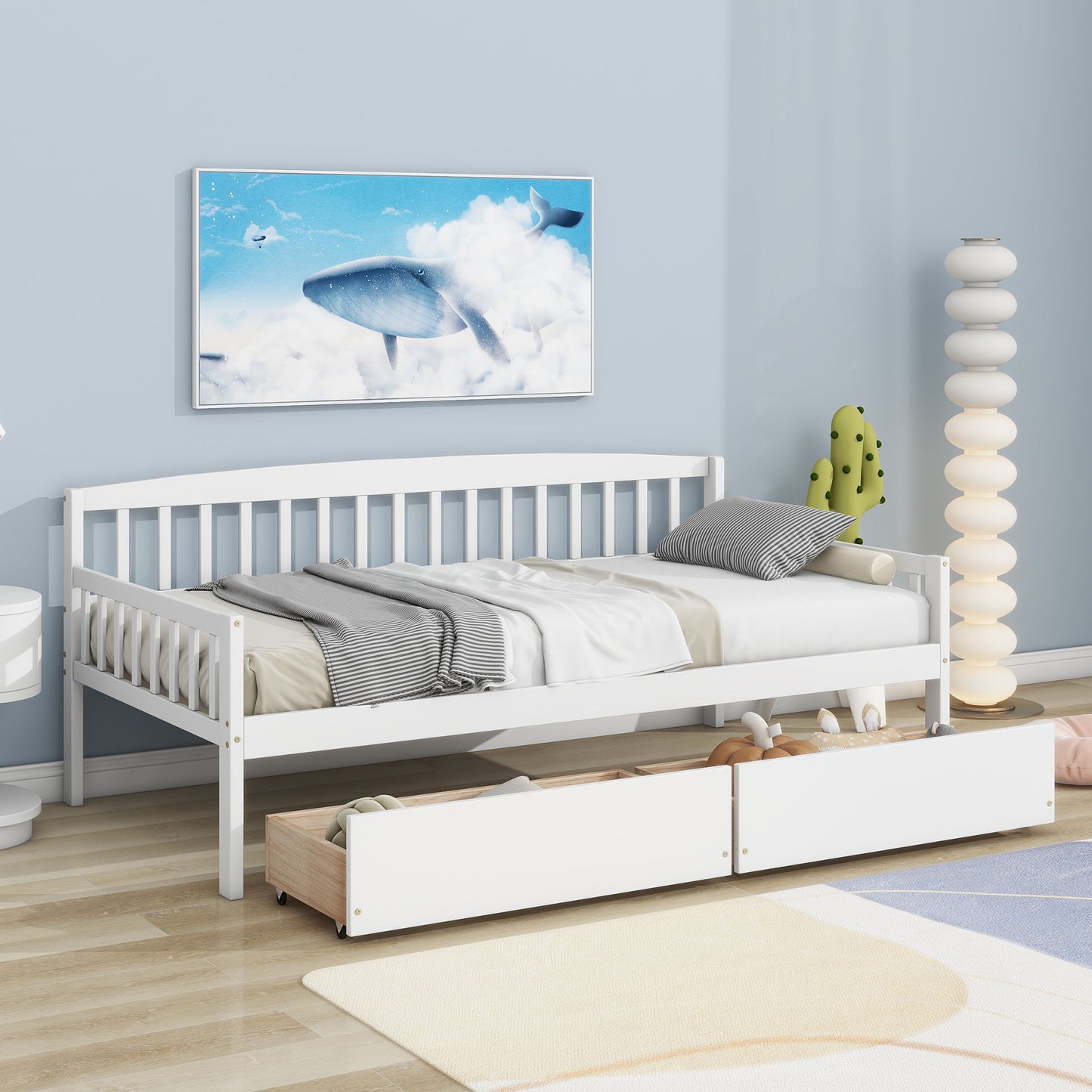 A Pine Wood Daybed With Two Storage Drawers and gray and white bedding sits against a blue wall, with two pull-out drawers below. Above is whale sky artwork, and a modern white lamp plus cactus decor are nearby.