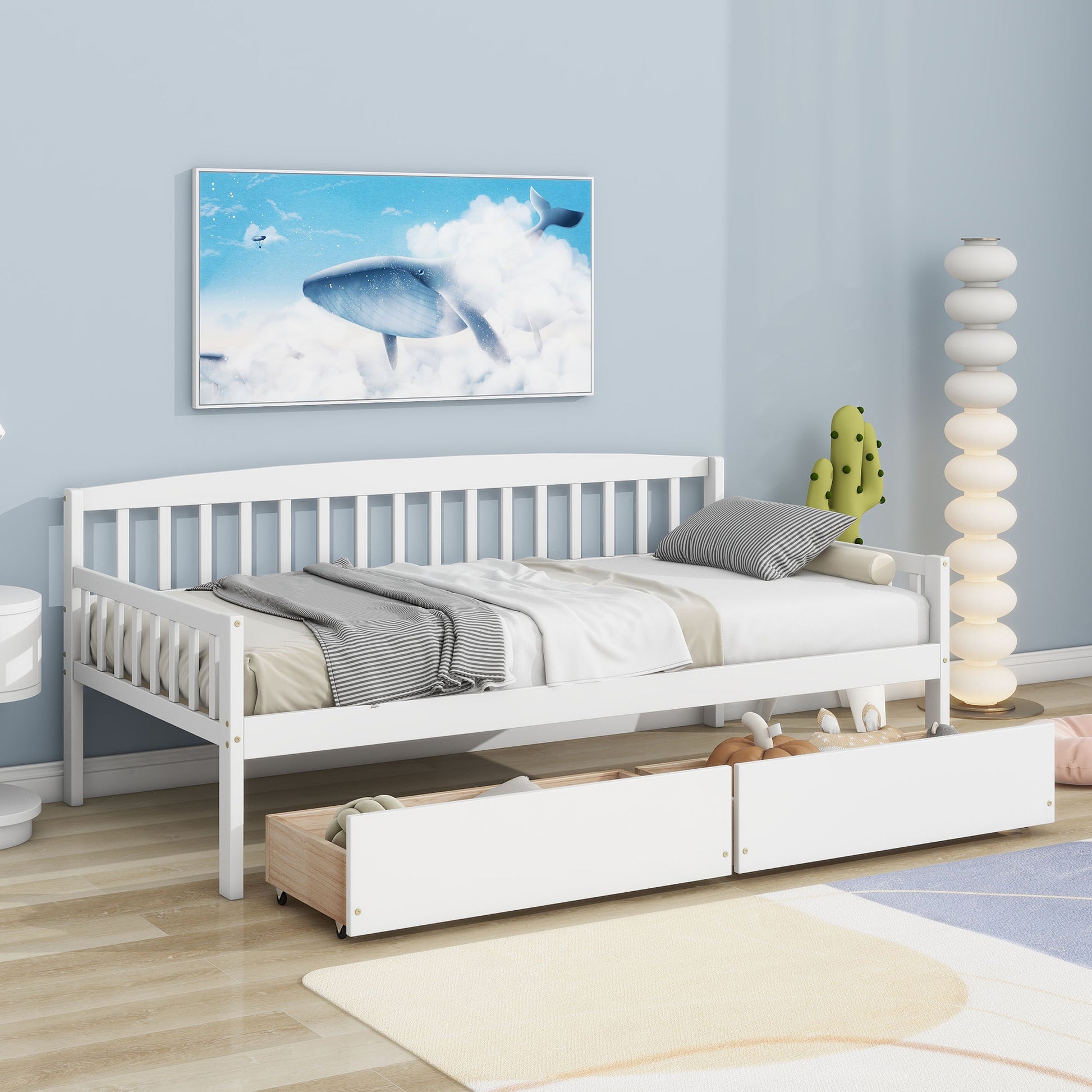 A Pine Wood Daybed With Two Storage Drawers and gray and white bedding sits against a blue wall, with two pull-out drawers below. Above is whale sky artwork, and a modern white lamp plus cactus decor are nearby.