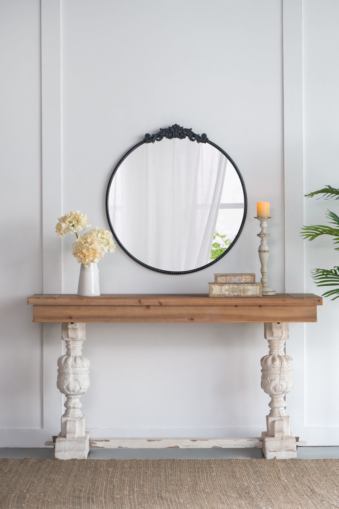 A rustic console table holds a vase of white flowers, books, and a candle. Above it hangs the Classic Design Mirror with Baroque Inspired Frame, leaning against a white paneled wall.