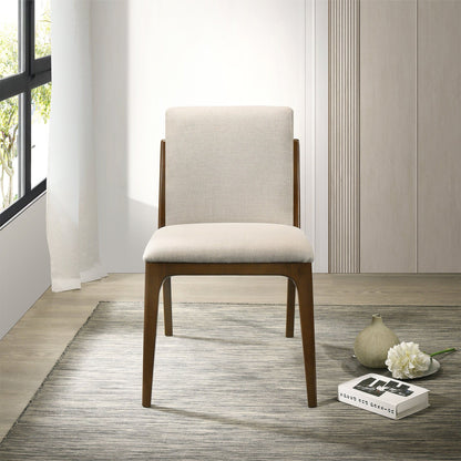 The Grivelle Elegant Dining Chair set, featuring modern wood design and light beige upholstery, stands on a gray rug near a gray vase, white flower, and two books. The space has light wood flooring, large windows, and neutral decor.