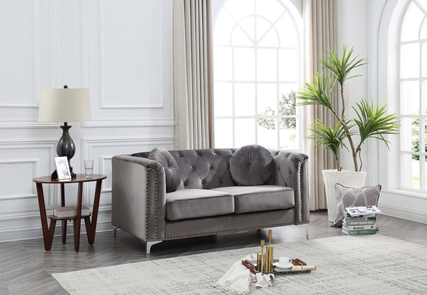 A bright living room features the Stylish Sloped Arm Loveseat in gray with round pillows, a tall potted plant, a lamp and books on a side table, cups on a tray atop a light rug, set before large arched windows with curtains.