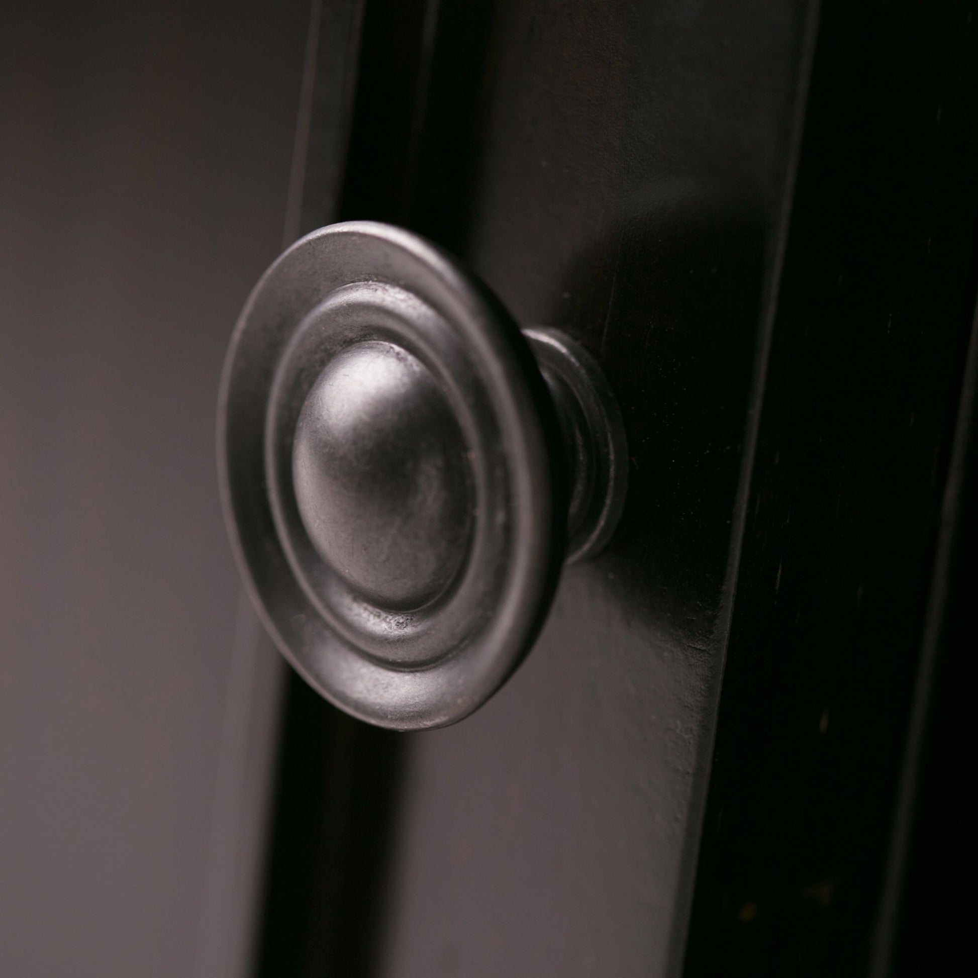Close-up of the Amherst - TV Media Stand, Handcrafted, showcasing its round metallic knob with a decorative circular design on a dark, smooth surface. Soft lighting highlights the elegant hardware detail.