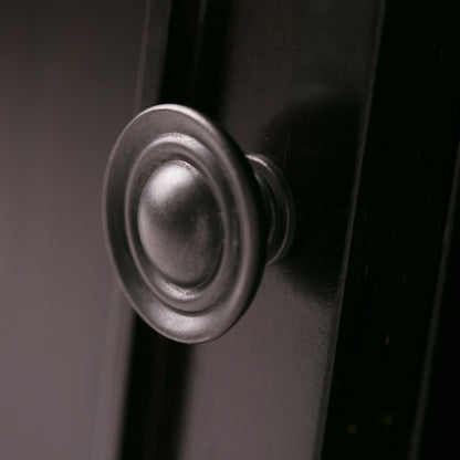 Close-up of the Amherst - TV Media Stand, Handcrafted, showcasing its round metallic knob with a decorative circular design on a dark, smooth surface. Soft lighting highlights the elegant hardware detail.