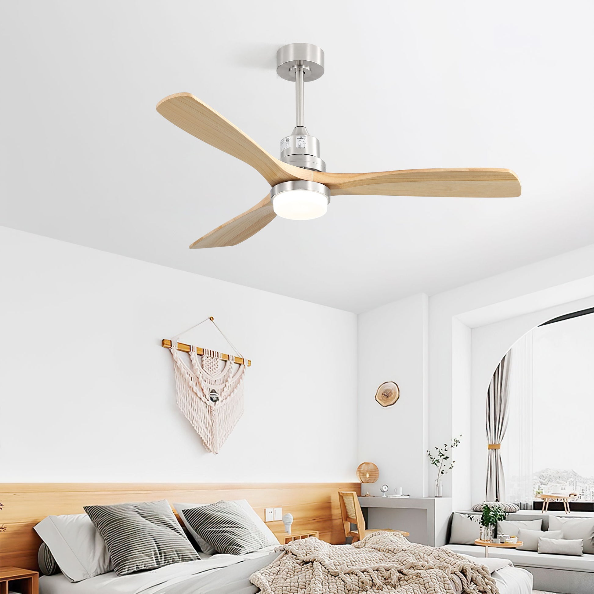 A modern bedroom features a wooden bed, cozy bedding, wall decor, and a brushed nickel ceiling fan with dimmable LED light, 3 wood blades, reversible DC motor, and 6-speed remote. Natural light streams through a large window with a sitting area.