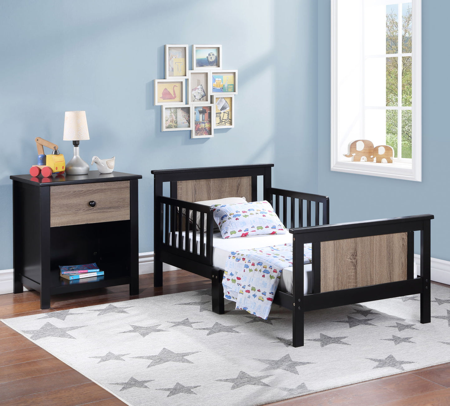 A toddler bedroom featuring the Connelly - Reversible Panel Toddler Bed in black and wood, colorful blanket, nightstand with a lamp and toys, wall art, large window on a light blue wall, and a gray star-patterned rug on the floor.
