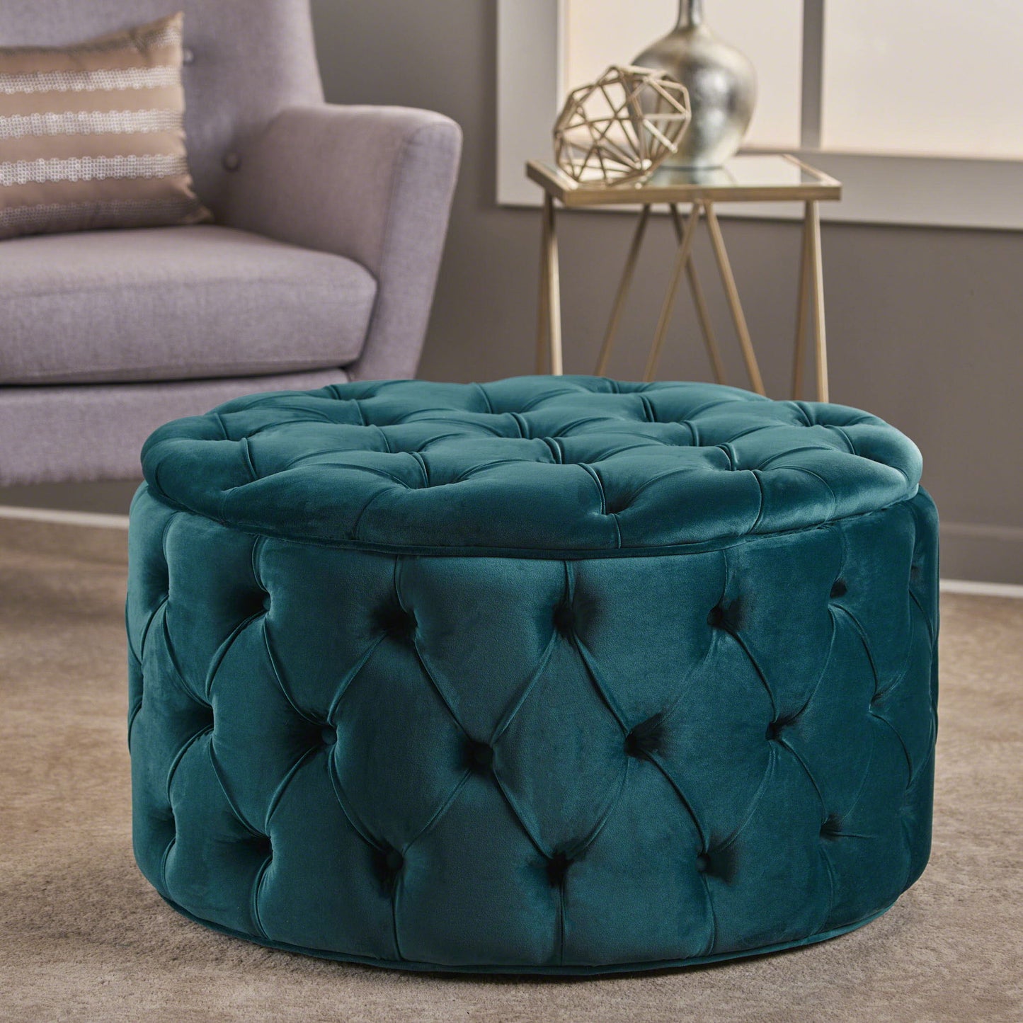 A Modern Glam Ottoman Velvet, round and teal with tufted details, sits on a beige carpet in a modern living room, alongside a gray armchair, side table, and decorative accents.