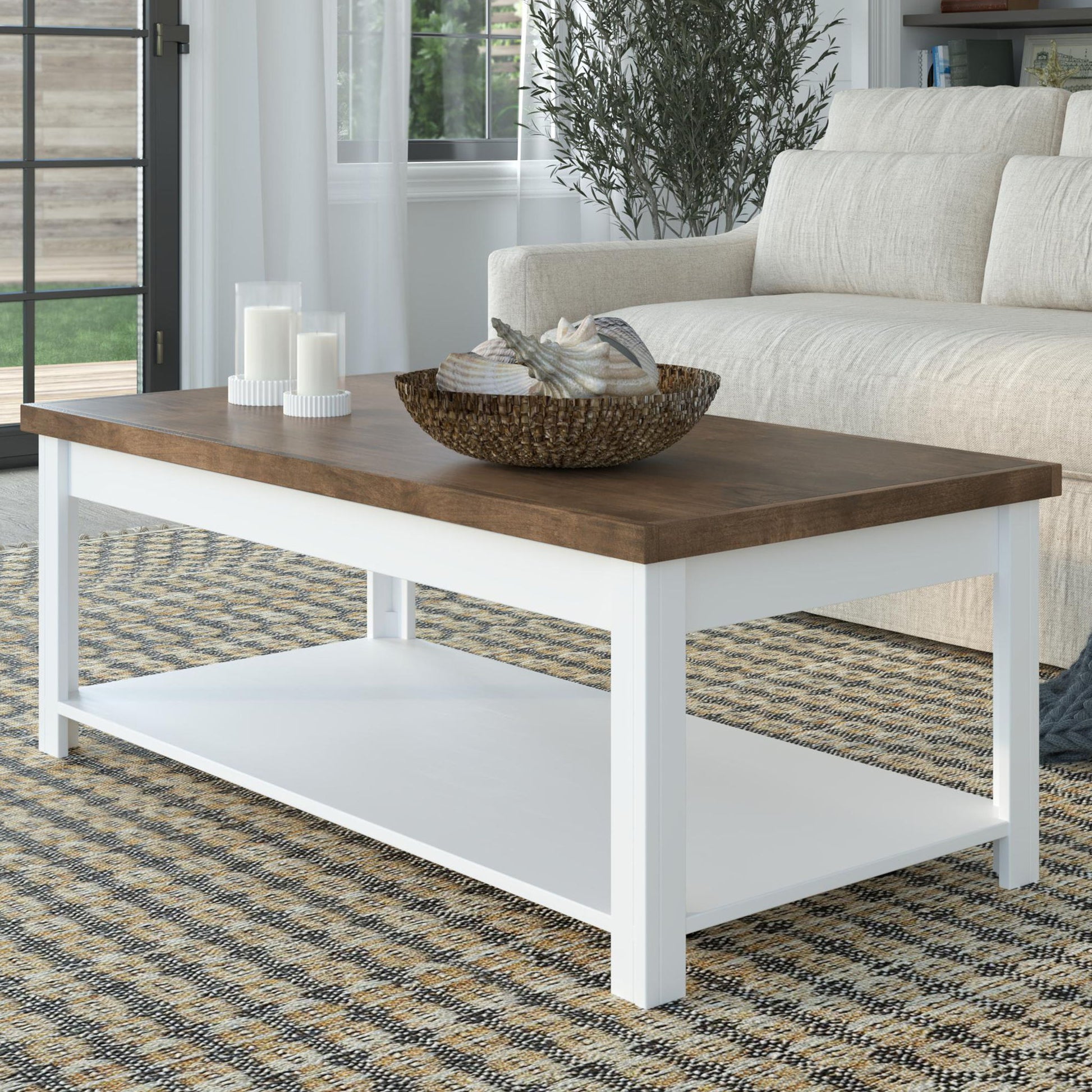 The Hampton - Coffee Table in Jasmine Whitewash/Barnwood sits on a patterned rug, displaying a basket of decor and three white candles. A beige sofa and green plant complete the living room setting.