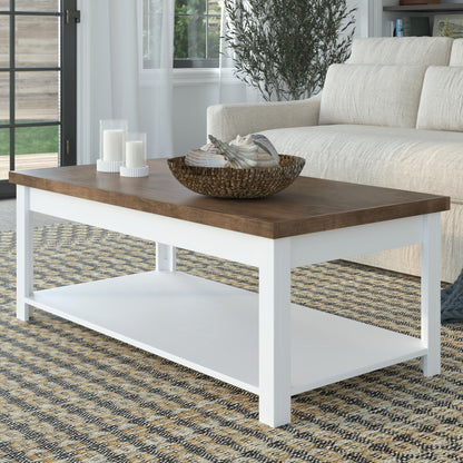 The Hampton - Coffee Table in Jasmine Whitewash/Barnwood sits on a patterned rug, displaying a basket of decor and three white candles. A beige sofa and green plant complete the living room setting.