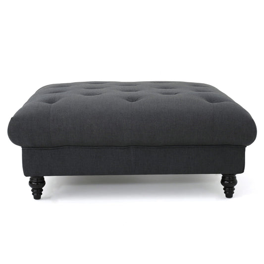 The Madrid - Tufted Ottoman Footrest is a dark gray, rectangular piece with a tufted top and four short, turned wooden legs, displayed against a white background.