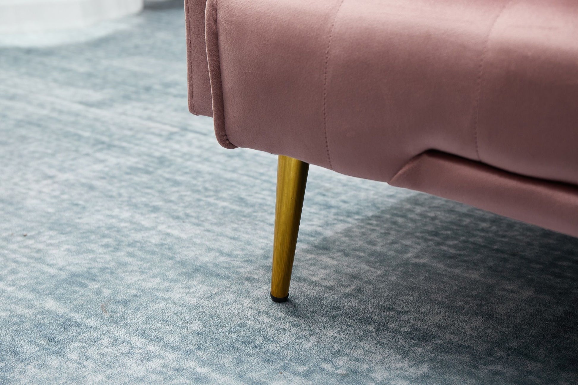 A close-up of the Comfort Compact Design Sofa Bed, featuring pink upholstery and gold metal legs, placed on a light blue textured carpet.