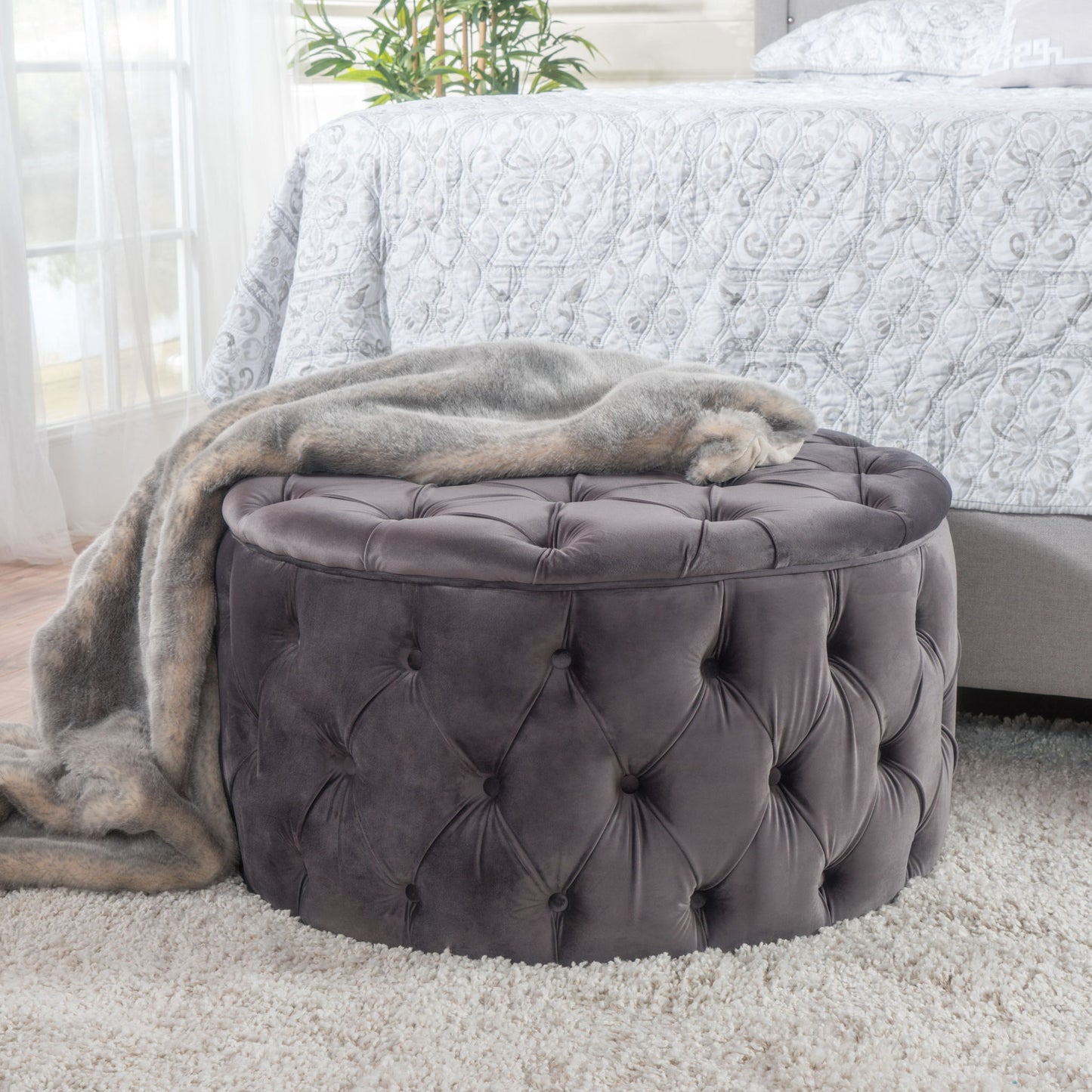A Modern Glam Ottoman Velvet in round tufted gray stands on a plush light carpet beside a bed with a patterned white comforter, draped with a soft gray faux fur throw. A plant decorates the background.