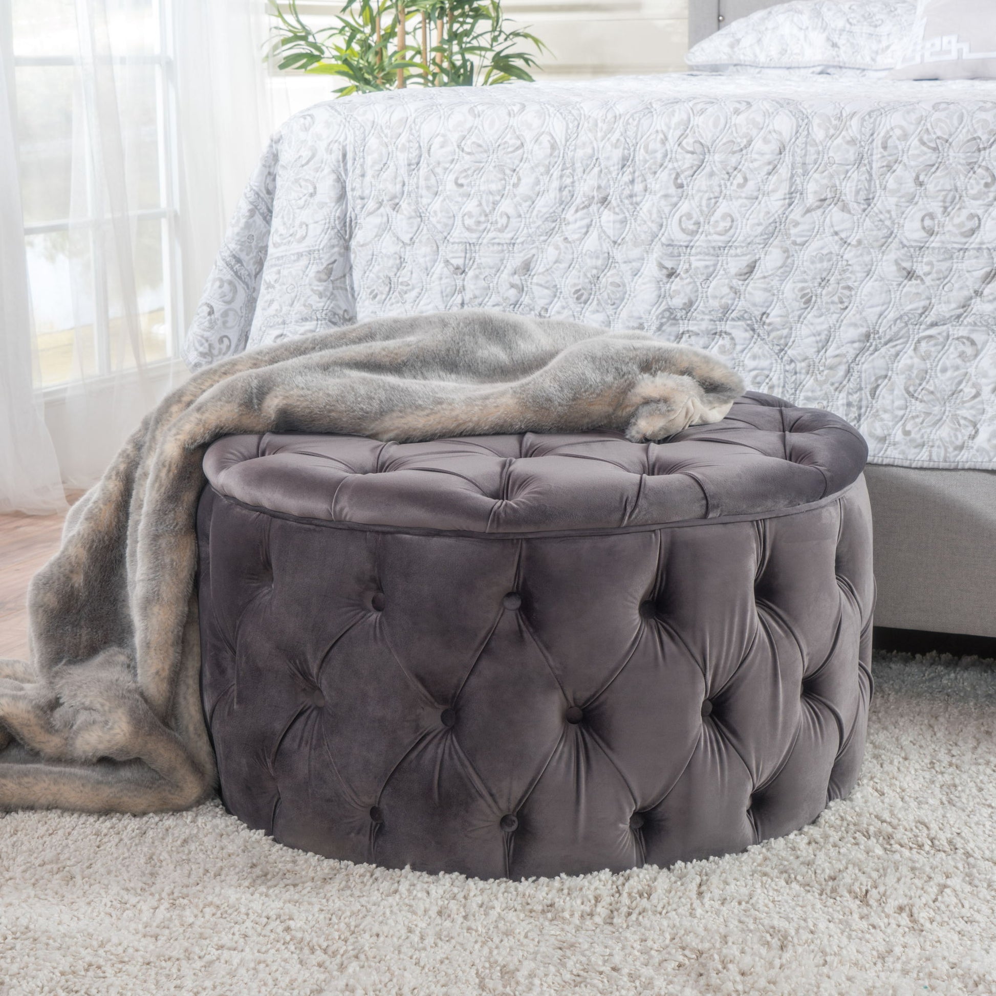 A Modern Glam Ottoman Velvet in round tufted gray stands on a plush light carpet beside a bed with a patterned white comforter, draped with a soft gray faux fur throw. A plant decorates the background.