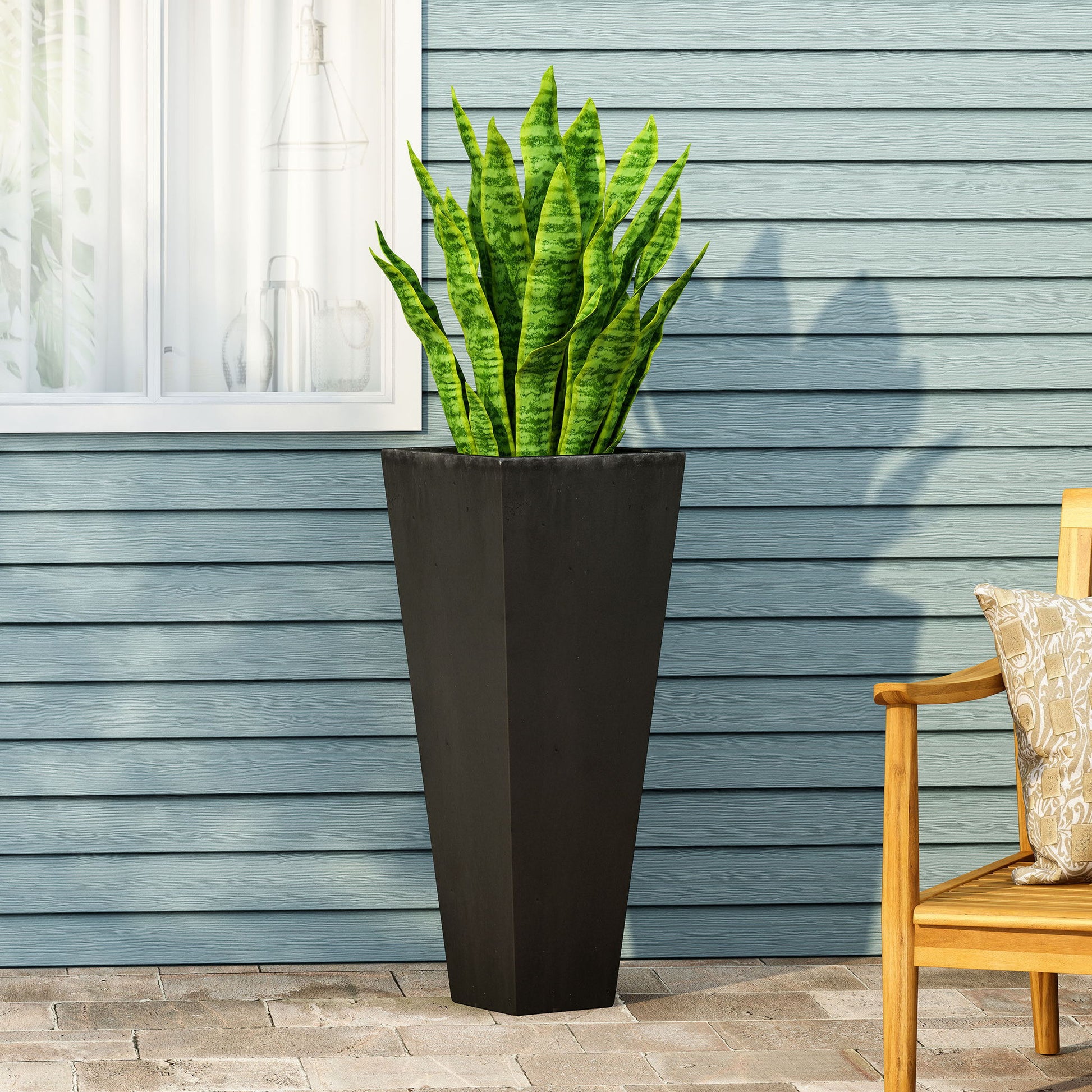 An Outdoor Modern Cast Stone Planter with a snake plant rests on a stone patio before a blue-gray paneled wall, beside a window and a wooden chair with a patterned cushion.