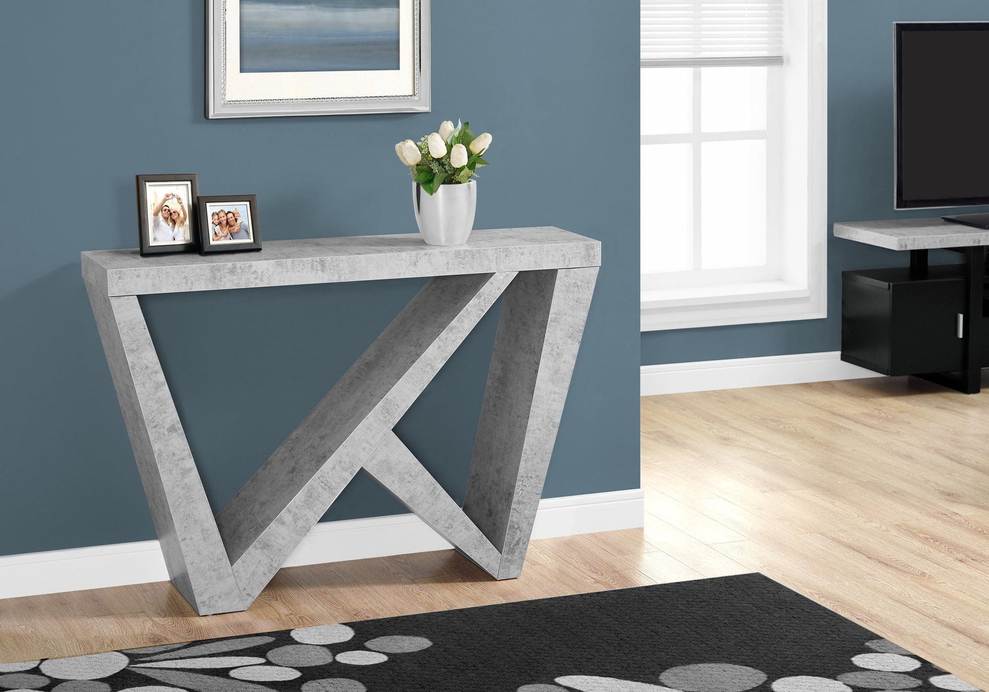 The Accent Console Table For Entryway with durable construction features a modern gray geometric design against a blue wall, topped with framed photos and a white floral vase. Light streams through the window, with a TV and rug partially visible.