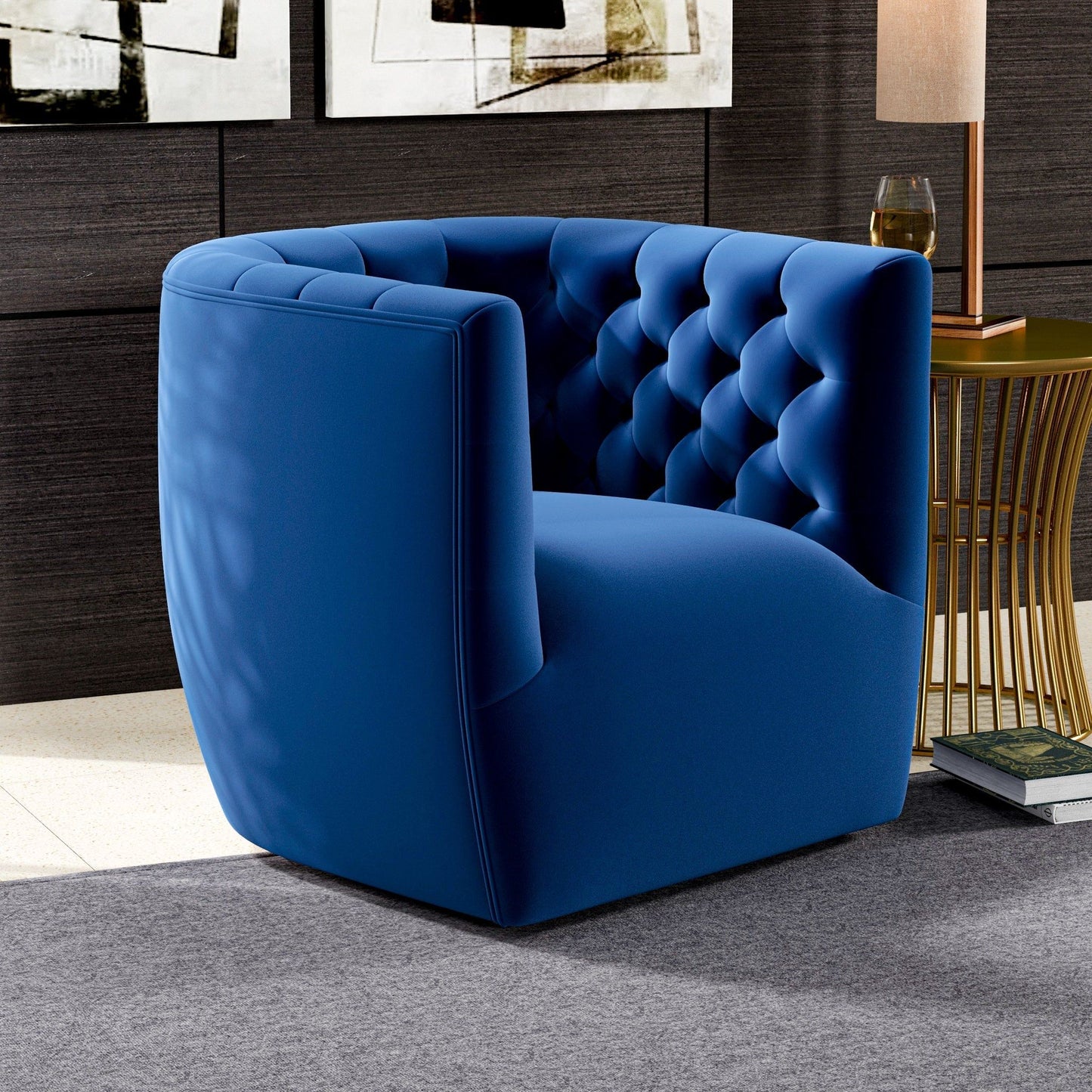 The Delaney Ergonomic Design Swivel Chair in blue velvet with a tufted backrest sits on a gray rug next to a gold side table with a lamp and glass, set in a chic room featuring dark wood walls and abstract art.