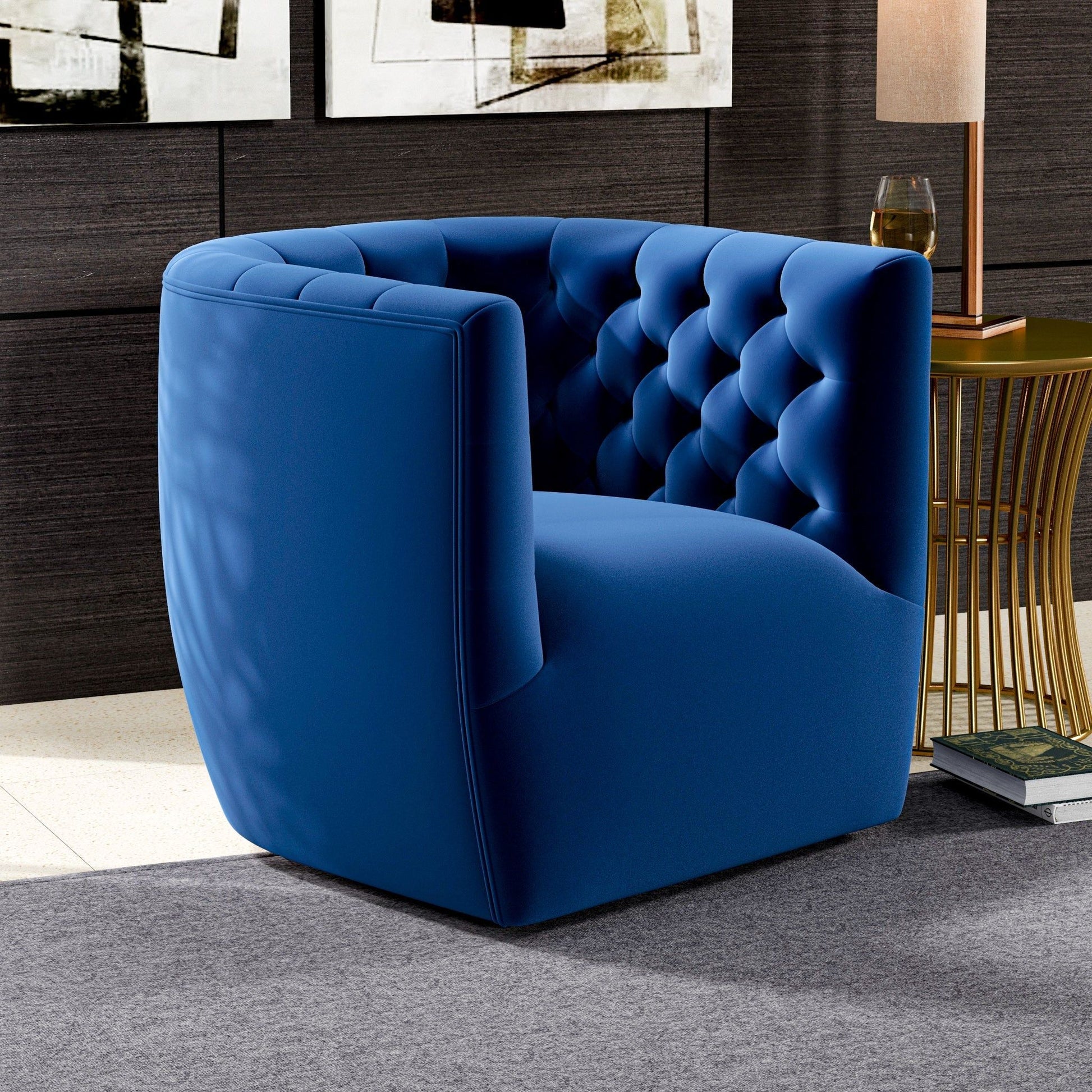The Delaney Ergonomic Design Swivel Chair in blue velvet with a tufted backrest sits on a gray rug next to a gold side table with a lamp and glass, set in a chic room featuring dark wood walls and abstract art.