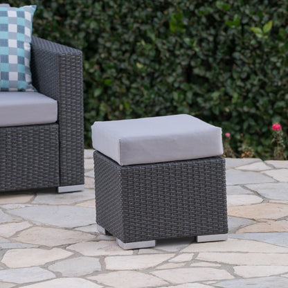 The Santa Rosa Outdoor Lounge Ottoman with a gray cushion is placed on a stone patio beside a matching wicker chair, with a green leafy hedge in the background.