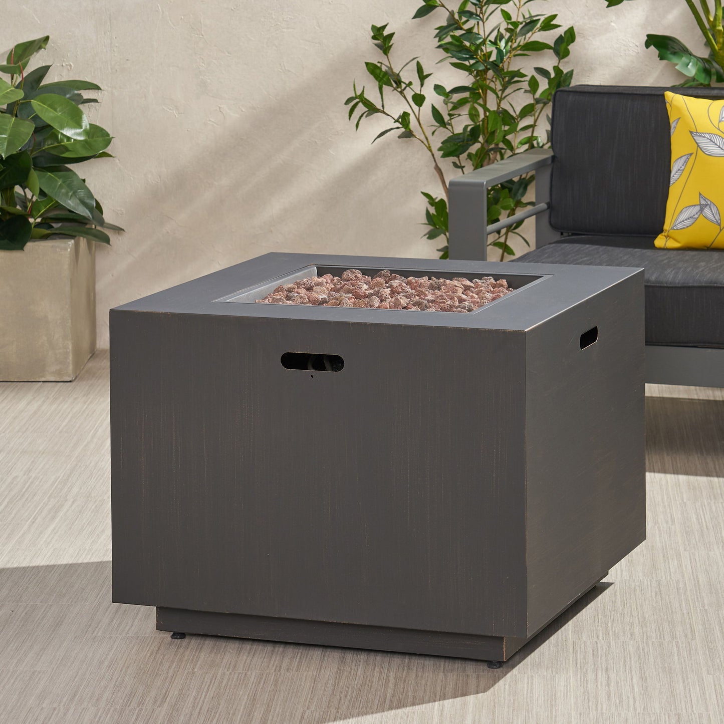 A square, dark gray Outdoor Patio Gas Burning Fire Pit Table 50,000 Btu sits on a light patio near a potted plant and a dark outdoor sofa with a yellow patterned pillow.