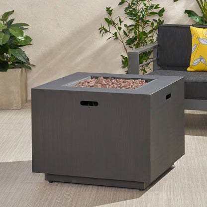 A square, dark gray Outdoor Patio Gas Burning Fire Pit Table 50,000 Btu sits on a light patio near a potted plant and a dark outdoor sofa with a yellow patterned pillow.