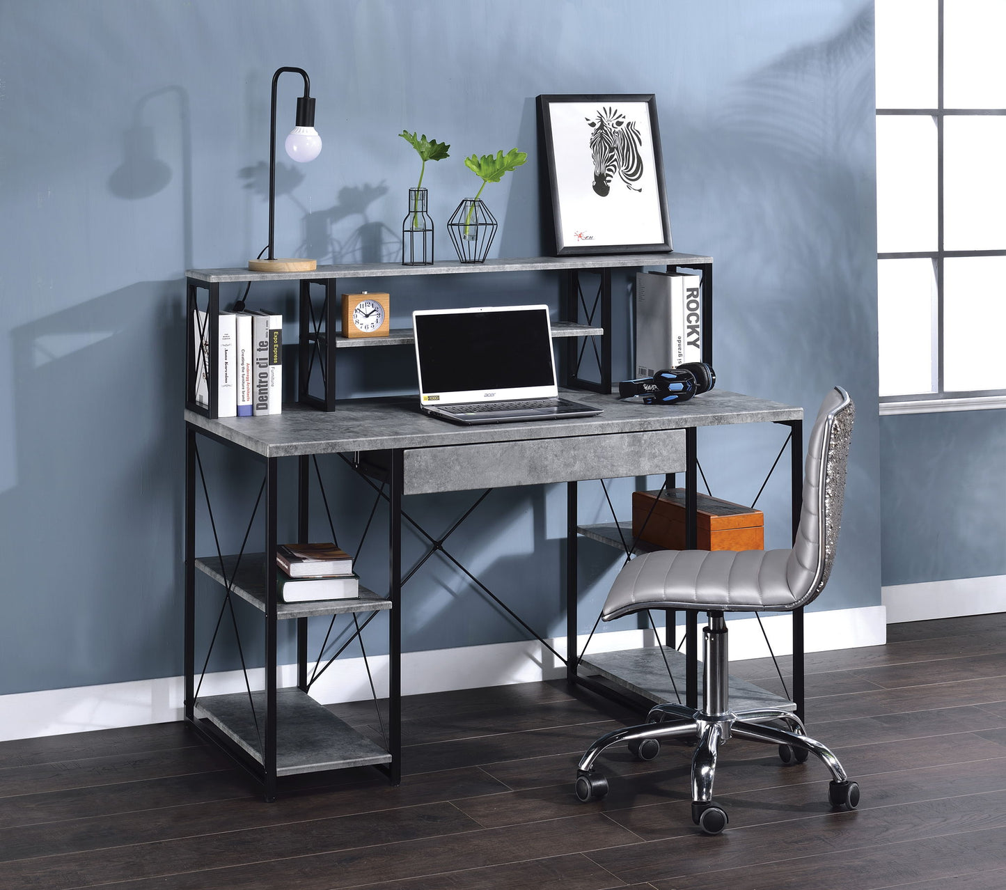 A sunlit, modern workspace featuring the Amiel - All Purpose Writing Desk, with a laptop, desk lamp, framed zebra art, small plants, books and a clock on shelves. A gray cushioned office chair sits in front of the desk by the window.