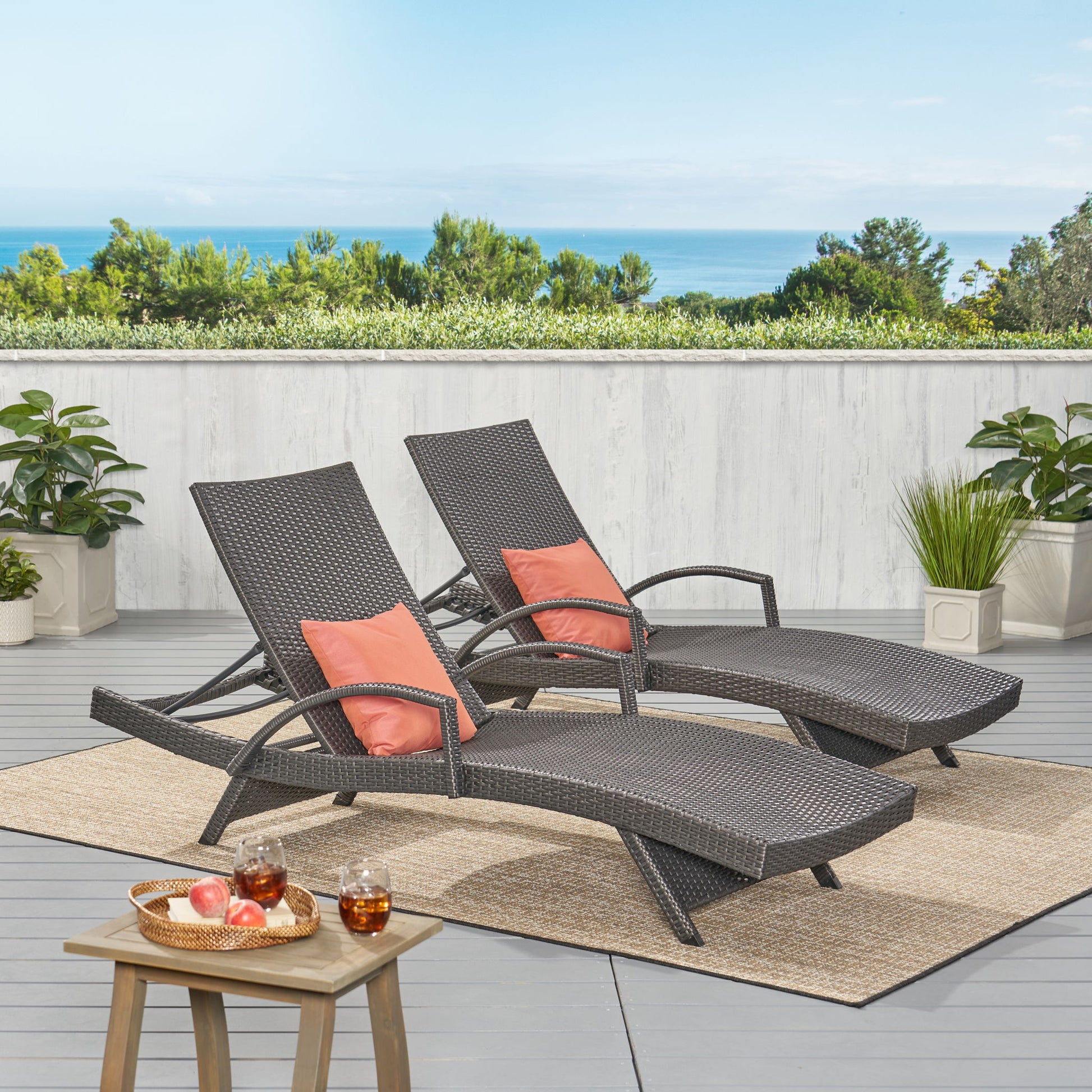 Two Salem - Contemporary Wicker Chaise Lounges with coral cushions sit on a patio rug facing lush greenery and an ocean view. A nearby wooden table holds drinks and apples, surrounded by potted plants.