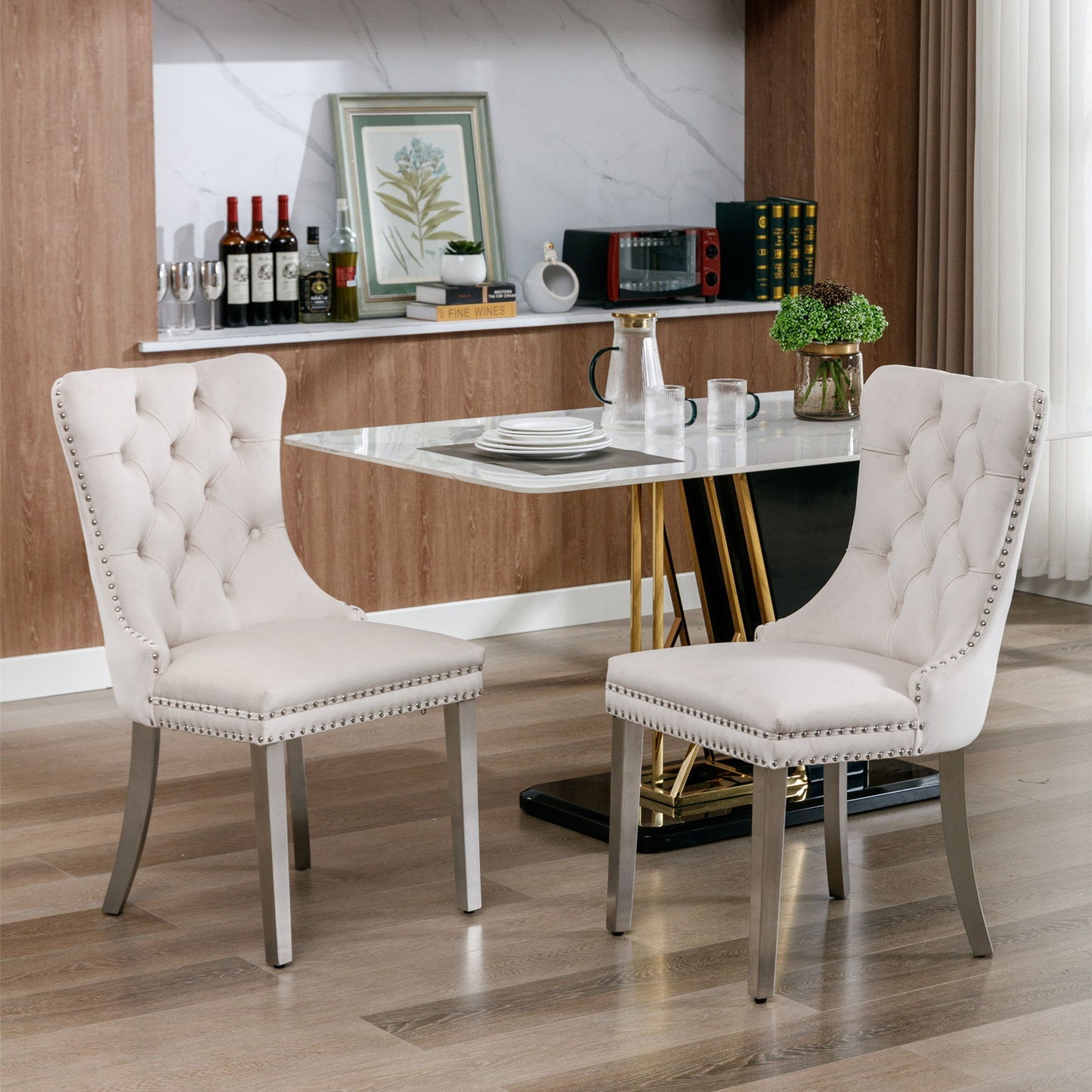 Two Nikki modern velvet upholstered dining chairs with tufted design, chrome stainless steel legs, and nailhead trim are placed before a marble dining table. Stylish kitchen shelves with bottles and decor can be seen behind them.