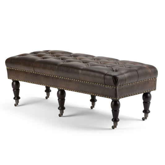 The Henley Tufted Ottoman Bench in distressed brown leather features brass nailhead trim and six turned wooden legs, each with a caster wheel at the base.