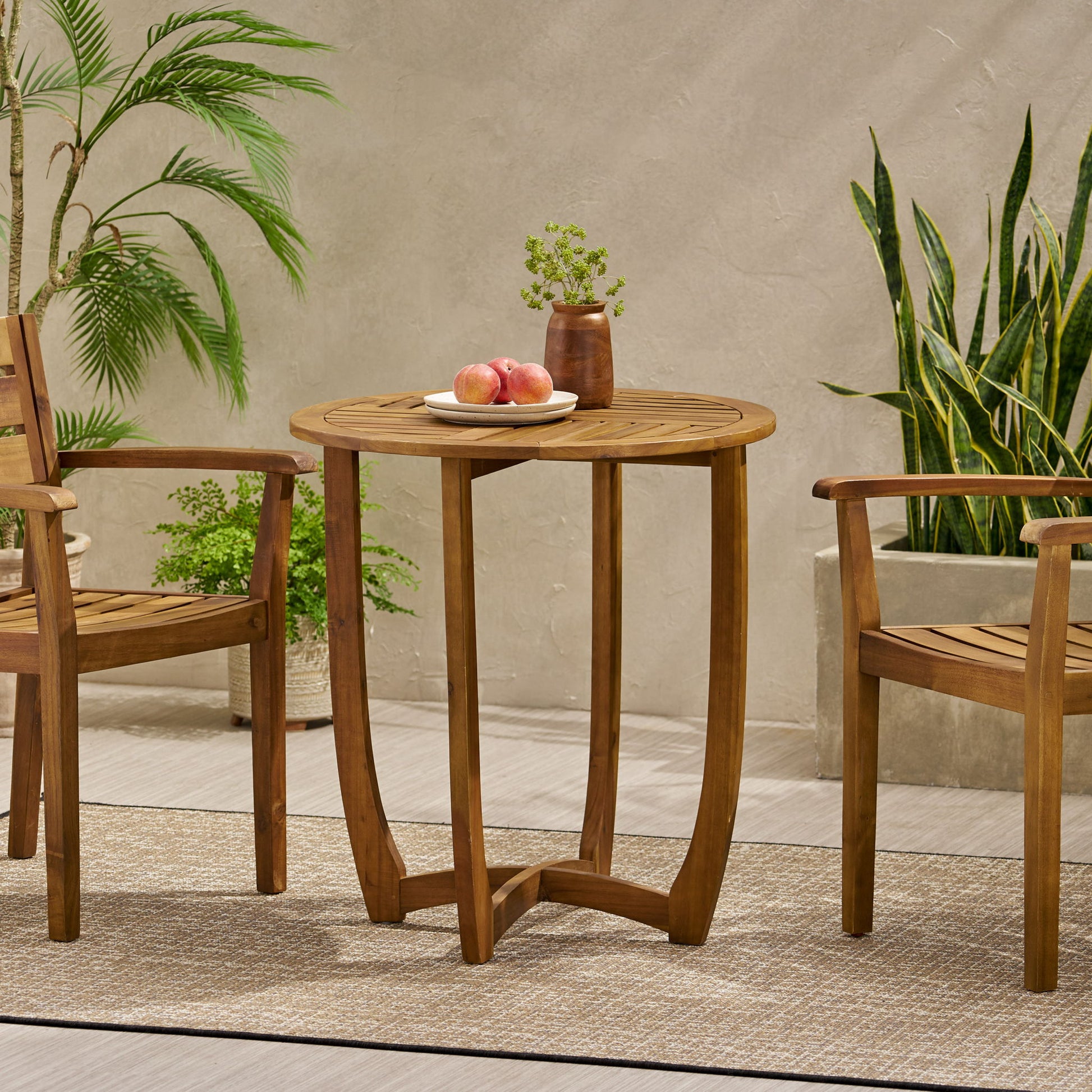 The Outdoor Circular Wood Bistro Table With Curved Legs And Slat Panel Table Top - Teak is set on a rug with two chairs. On the table are a plate of peaches and a brown vase with greenery. Potted plants line the beige wall behind.