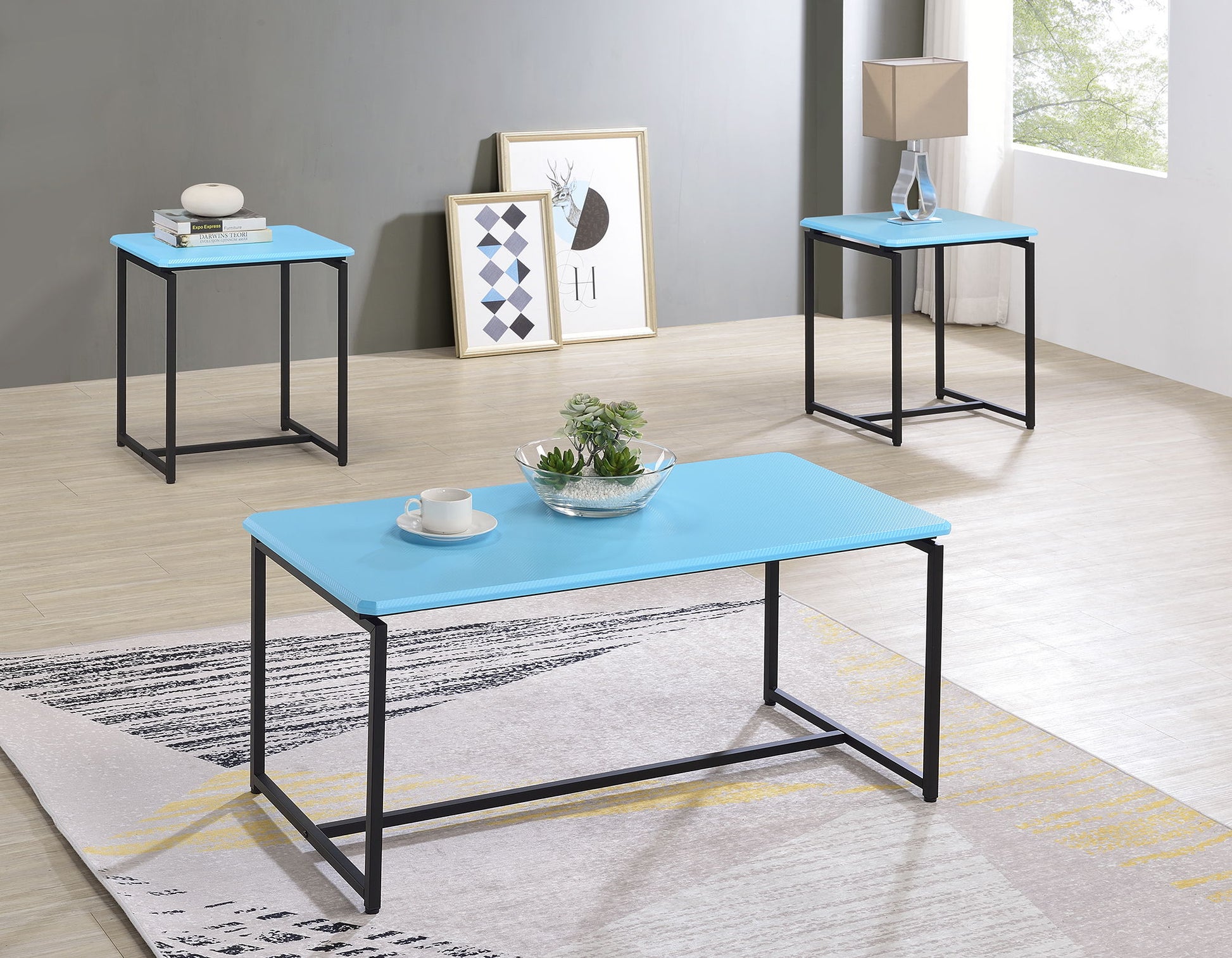 The GT - 3 Piece Carbon Fiber Wrap Coffee Table And End Table set features modern blue tops and black metal legs, arranged in a bright room with light wood floors, accented by a lamp, books, decor, framed art, and natural light from a large window.