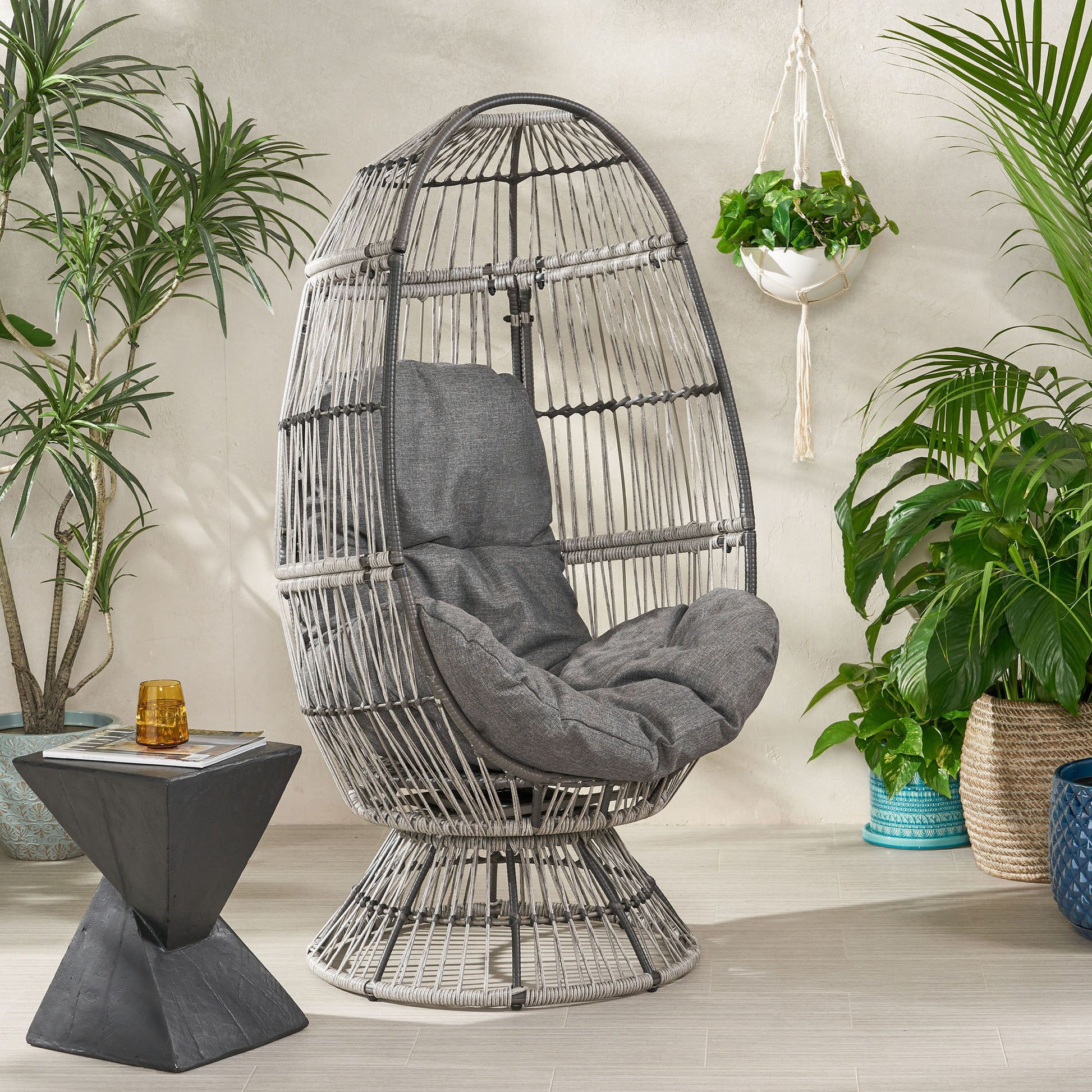The Patio Swivel Basket Egg Chair With Cushion, featuring gray cushions, sits between green potted plants. Nearby is a small geometric side table with an amber glass, all arranged on a light tile floor against a beige wall.