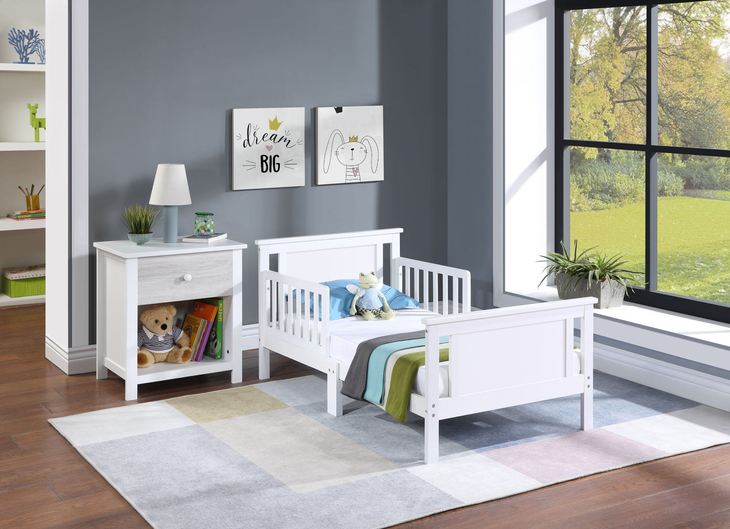 A tidy children’s bedroom features the Connelly Reversible Panel Toddler Bed in white with stuffed toys and a striped blanket, next to a white nightstand holding a lamp and books. Framed animal prints hang on the wall near a large garden-facing window.