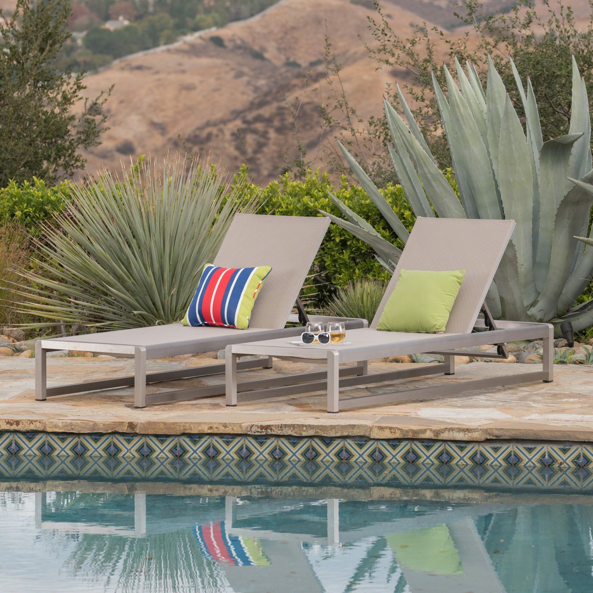 Two Outdoor Patio Chaise Lounges with 5 reclining positions and colorful pillows sit by a tiled pool. A small table between them holds sunglasses and a drink, with large agave plants and hills in the background.