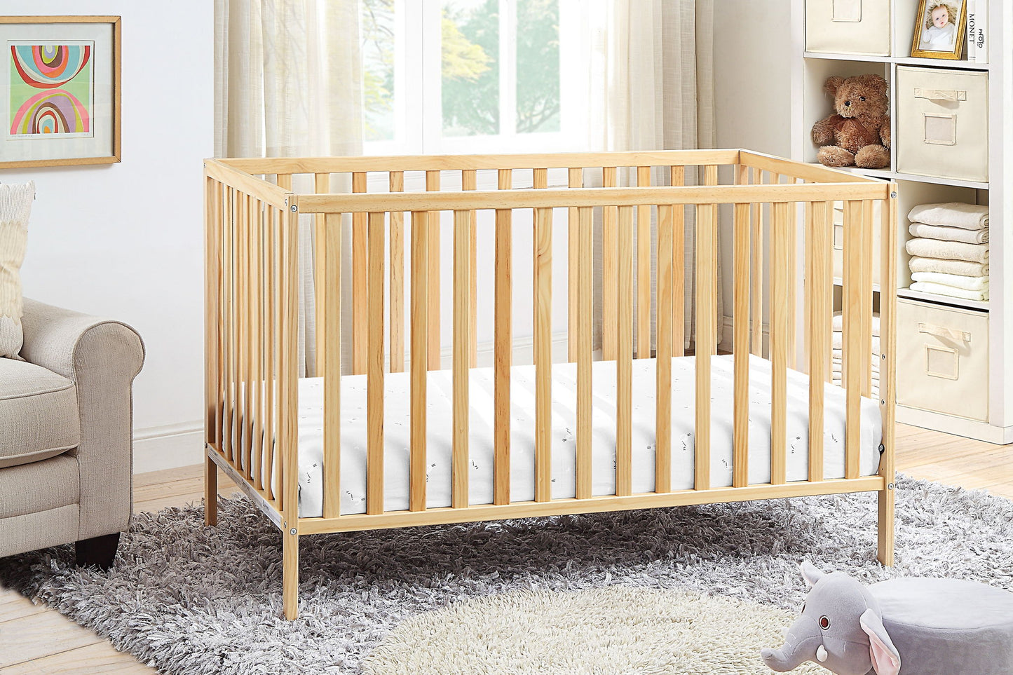 The Palmer - 3-in-1 Convertible Island Crib with a white mattress stands on a gray rug in a bright nursery. Nearby shelves hold folded linens and a teddy bear, while a plush gray elephant toy rests on the floor.