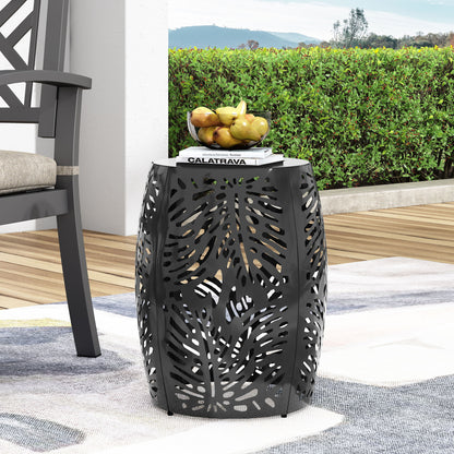 The Modern Design Metal End Table, Large, in black metal with a cutout leaf pattern, stands on a patio holding a stack of books and a bowl of fruit. Green hedges and distant hills can be seen in the background.