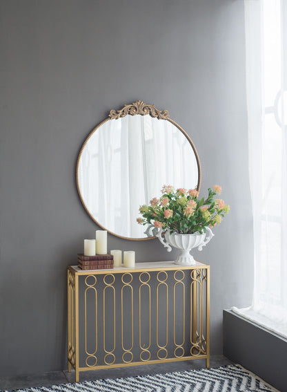 A Classic Design Mirror with a Baroque-inspired frame hangs above a gold console, styled with white candles, stacked books, and a vase of pink and green flowers. Sheer curtains filter soft natural light from the window.