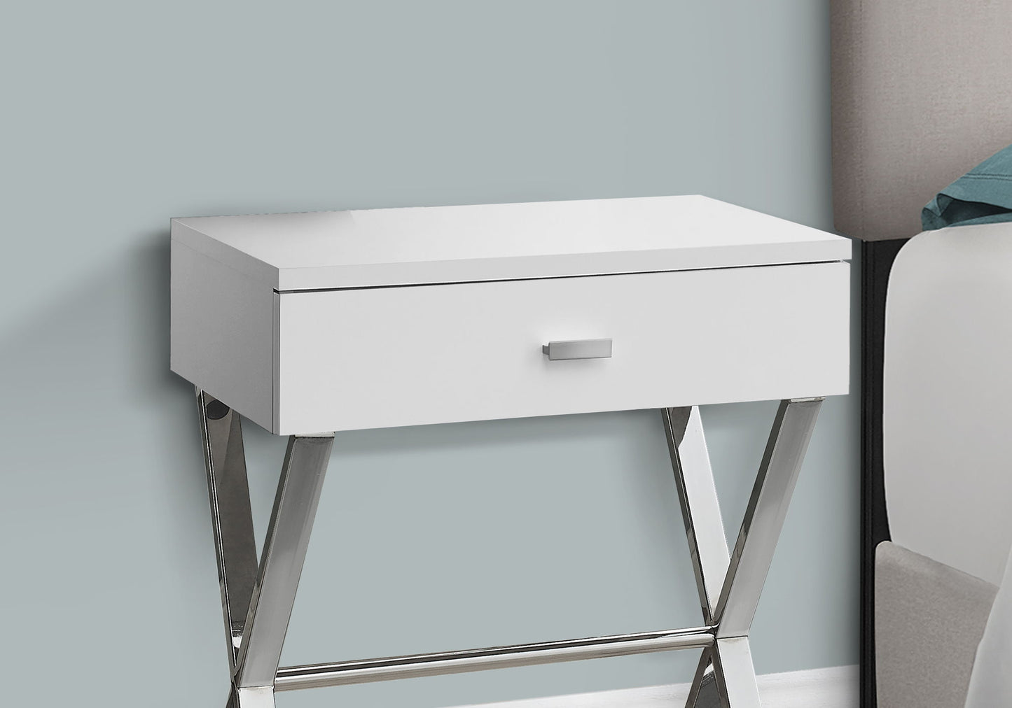 The Accent Side X Table, featuring a storage drawer and contemporary design, stands against a light blue wall next to a bed.