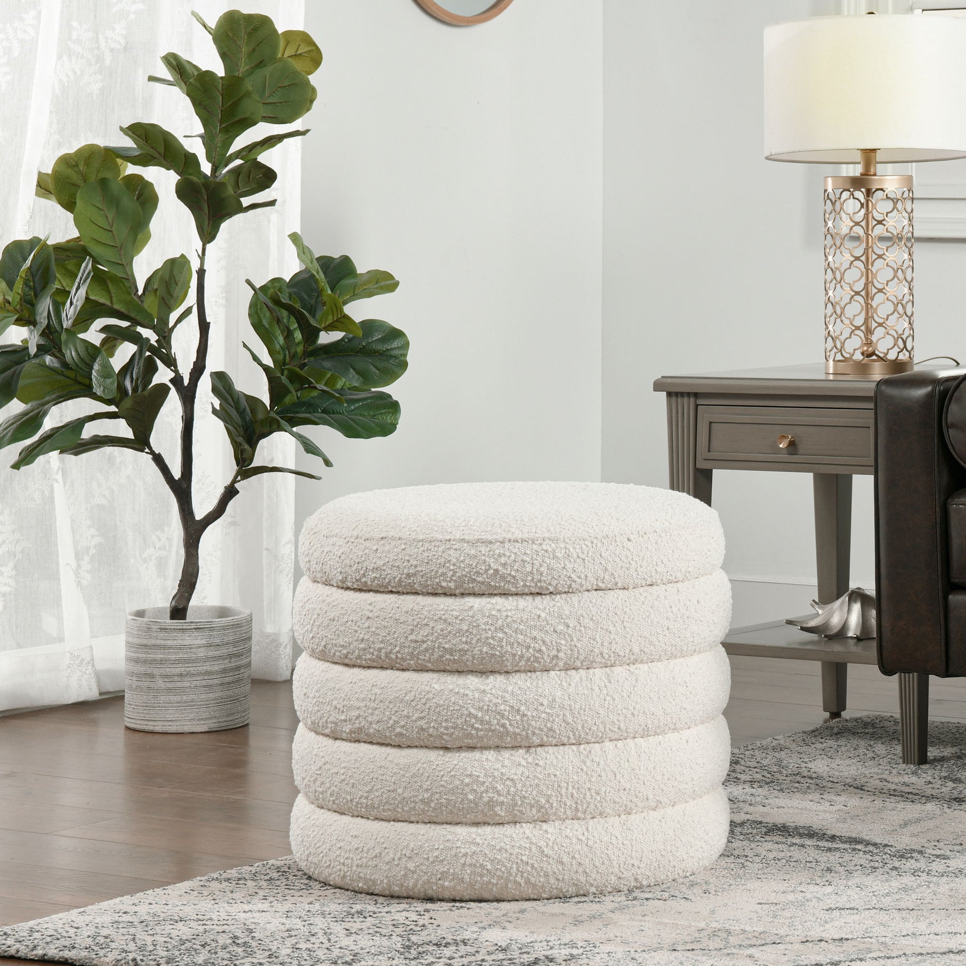 The Fuji Oversized Round Storage Ottoman, featuring a plush white design with stacked rings, sits on a patterned rug in a modern living room beside a dark wood side table and a potted plant near sheer-curtained windows.