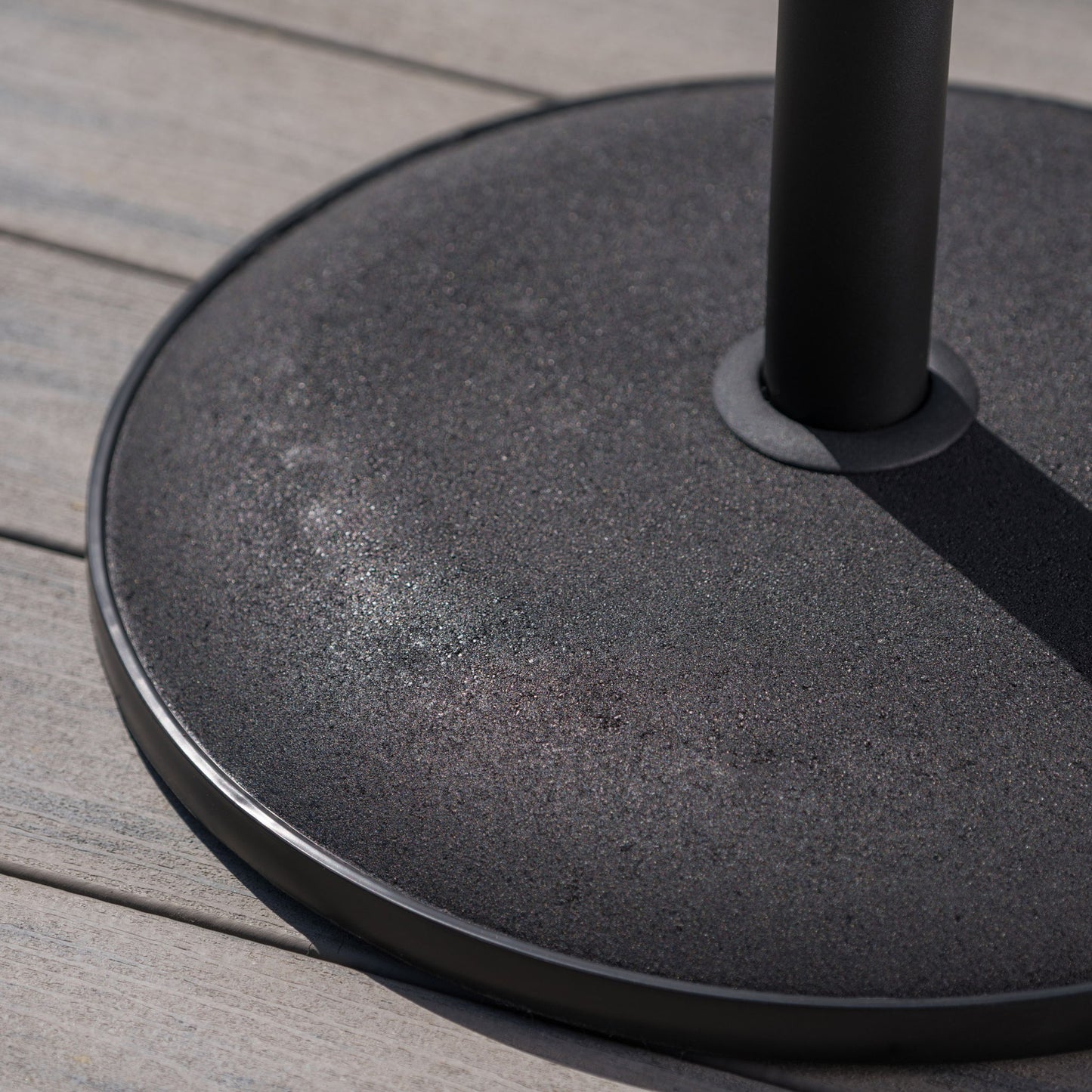 A close-up of the Stylish Concrete Umbrella Base With Iron Support Pole - Black, featuring its round, textured design as it rests on light gray wooden decking, with sunlight highlighting subtle reflections on its surface.