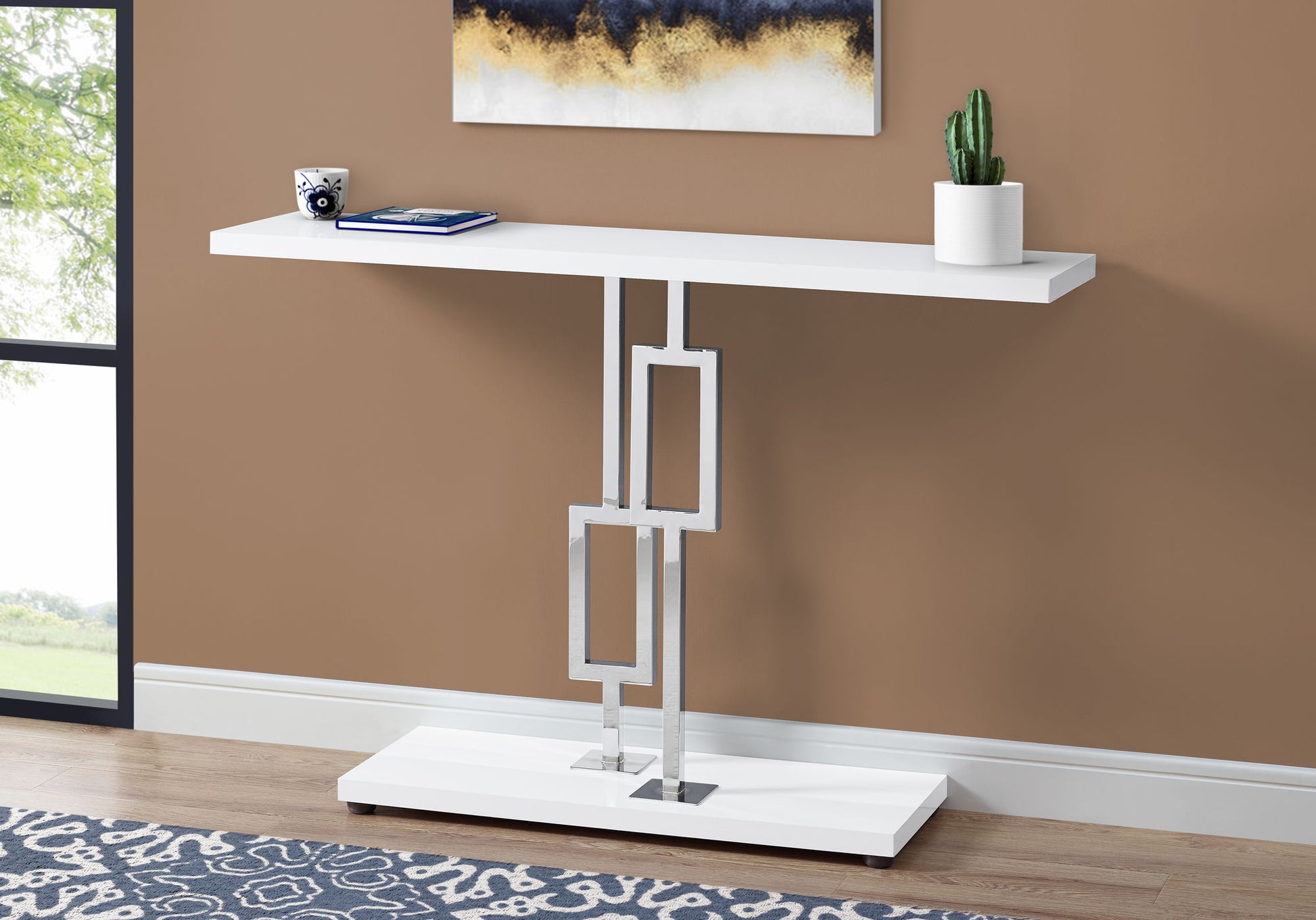 A Console Accent Table, Narrow For Living Room, featuring geometric silver legs and a white finish, is placed against a brown wall under abstract art, adorned with a camera, closed notebook, and potted cactus.