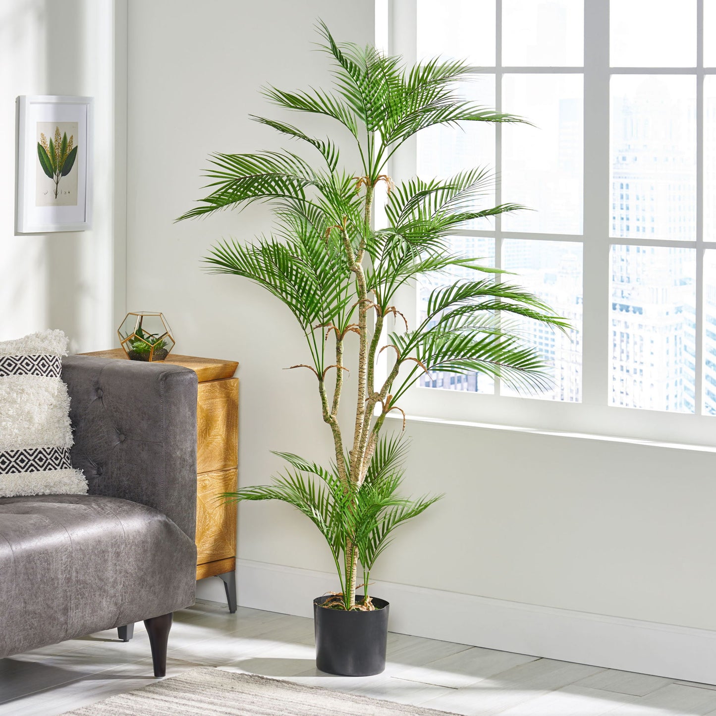 An Artificial Palm Tree stands next to a gray sofa in a bright living room with large windows, light walls, modern decor, a small wooden dresser, and framed artwork.