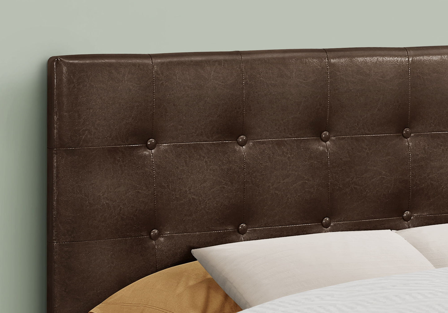 Close-up of the Full Size Upholstered Transitional Bed Headboard in brown, featuring tufted detailing and button accents behind bed pillows. A soft, muted green wall serves as the backdrop.