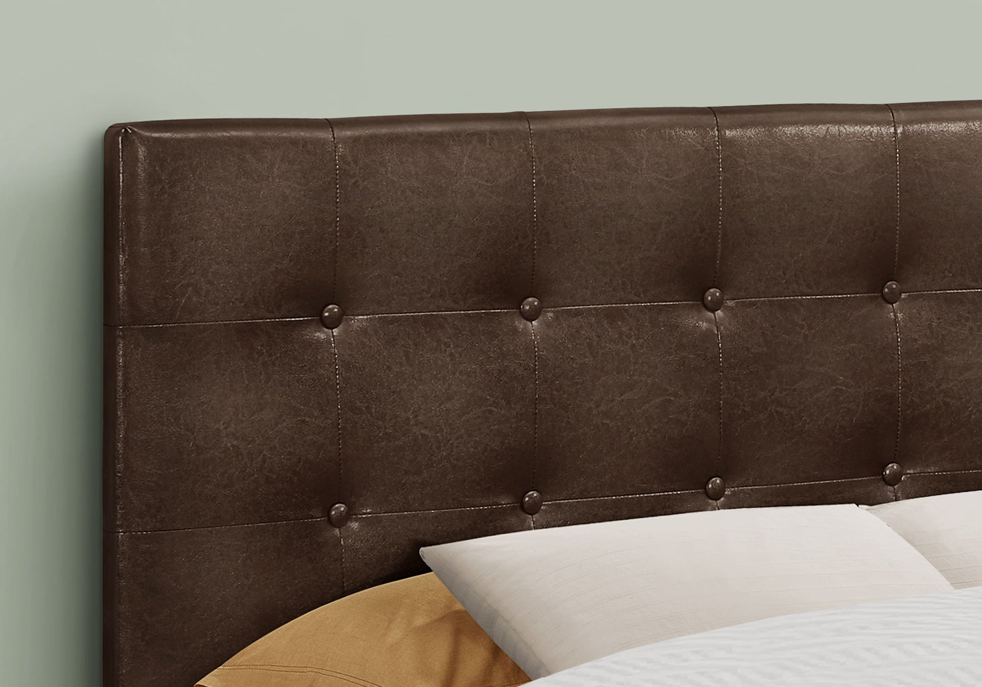 Close-up of the Full Size Upholstered Transitional Bed Headboard in brown, featuring tufted detailing and button accents behind bed pillows. A soft, muted green wall serves as the backdrop.