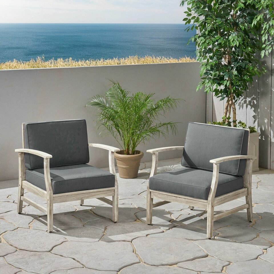 Two Perla - Contemporary Club Chairs with water resistant cushions sit on a stone patio with potted plants. A low wall is behind them, and beyond it, the ocean stretches out under a clear sky.
