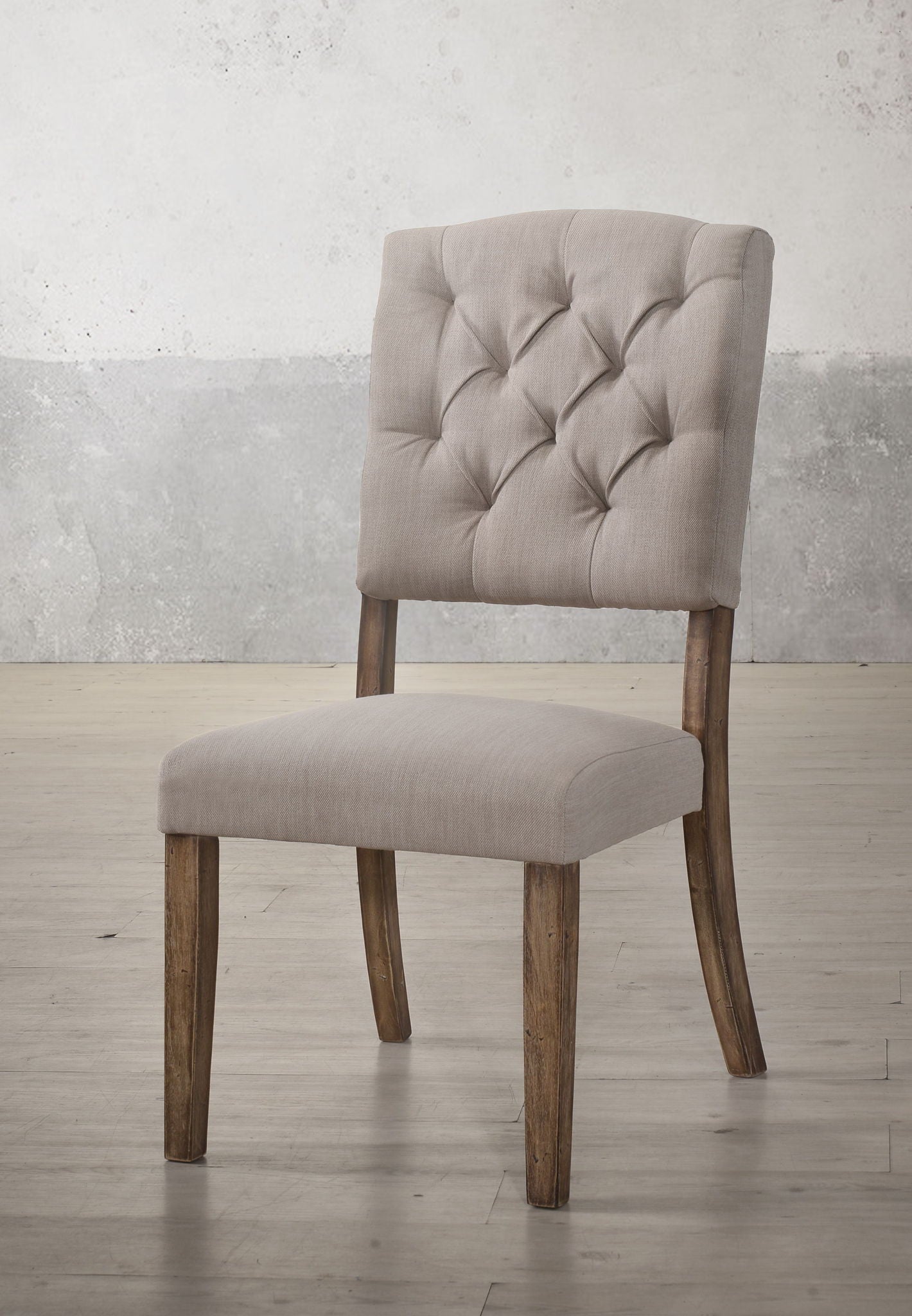 The Bernard - Weathered Side Chair (Set of 2) features beige upholstery, a tufted backrest, and wooden legs, shown on a light wood floor against a textured light gray wall.