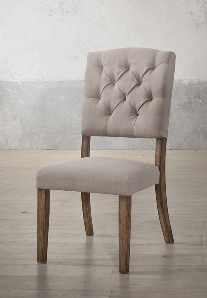 The Bernard - Weathered Side Chair (Set of 2) features beige upholstery, a tufted backrest, and wooden legs, shown on a light wood floor against a textured light gray wall.