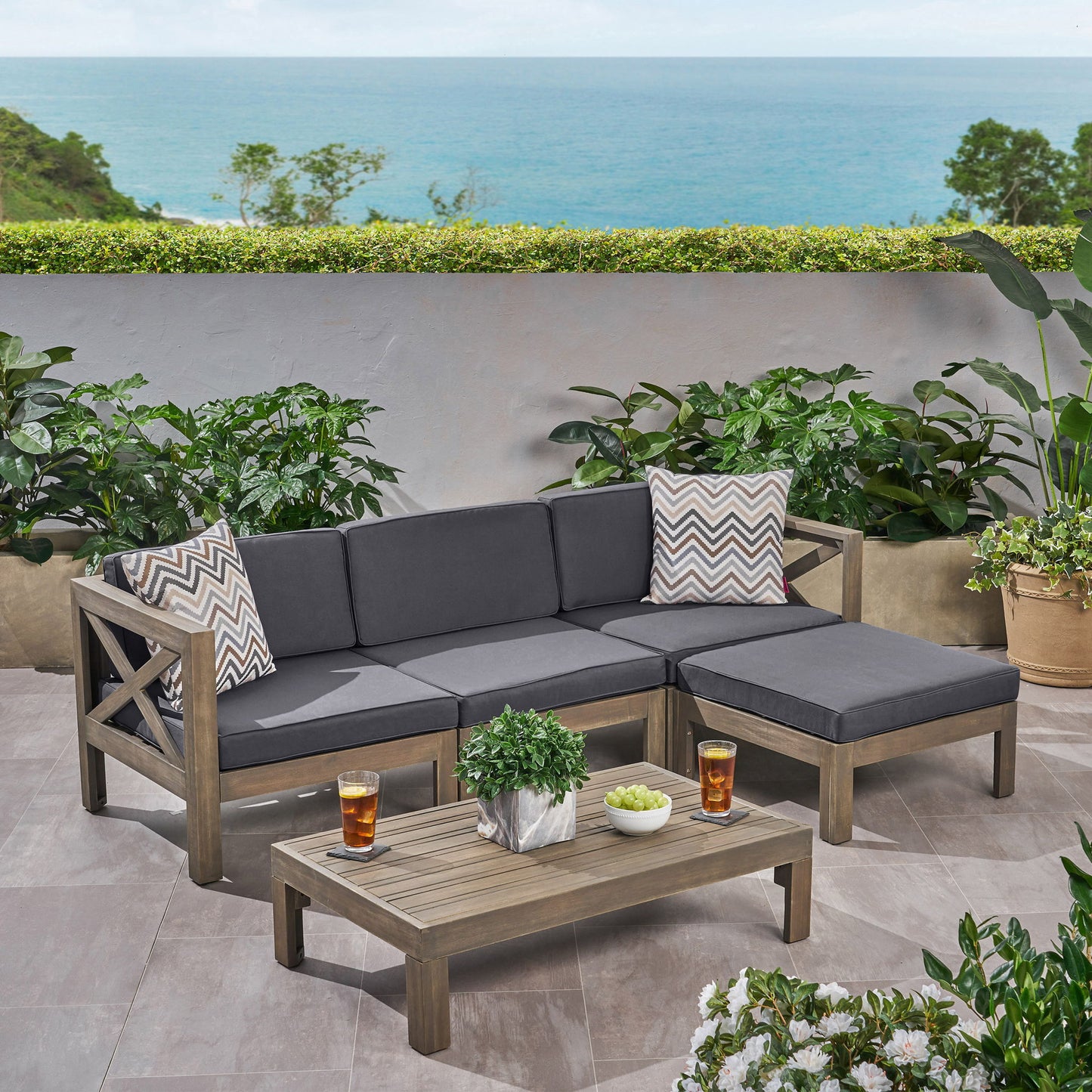 The Alcove L Shape Sofa Set features gray cushions, chevron-patterned pillows, and a wooden coffee table with drinks and plants, set amid greenery with an ocean view.
