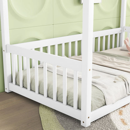 A close-up of the Canopy Frame Floor Bed With Fence, Guardrails in white wood with vertical slats, on a light wooden floor beside a green textured wall and patterned rug. Blankets and a stuffed toy rest on the bed.