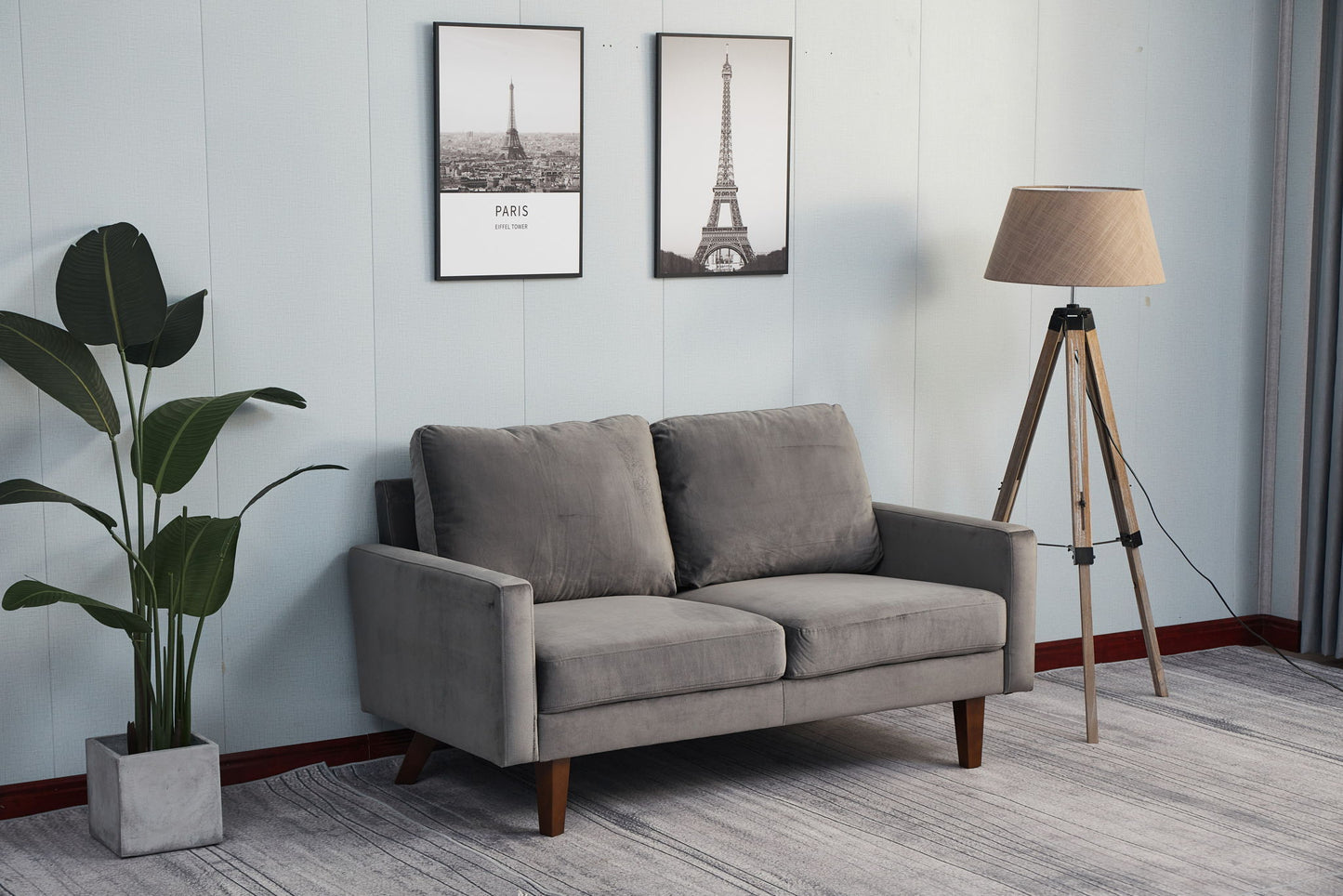 A modern living room featuring the Comfortable Loveseat With Durable Construction in gray, a tall potted plant, a tripod floor lamp, and two framed Eiffel Tower pictures on a light-colored wall.
