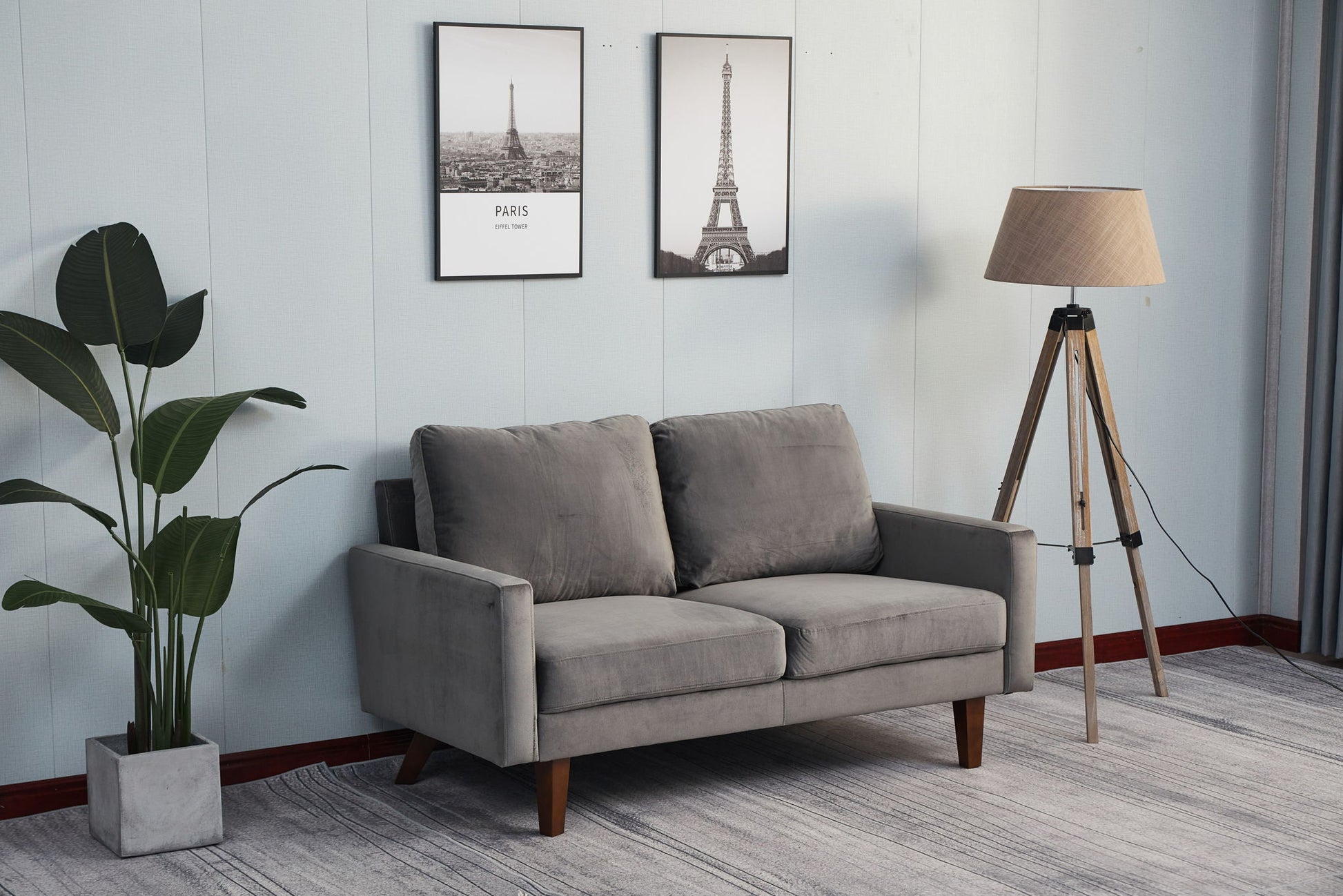 A modern living room featuring the Comfortable Loveseat With Durable Construction in gray, a tall potted plant, a tripod floor lamp, and two framed Eiffel Tower pictures on a light-colored wall.