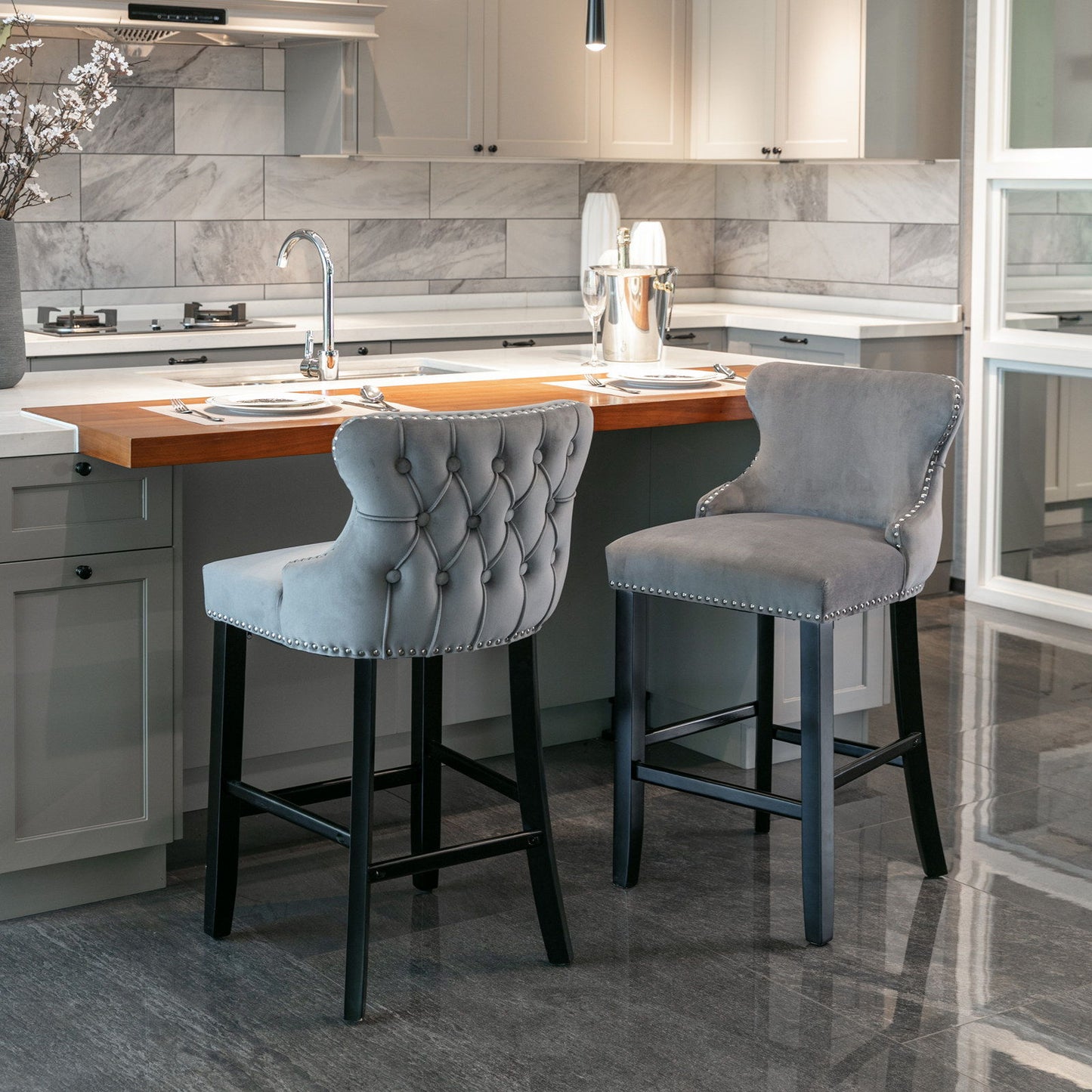 Two Contemporary Velvet Upholstered Wing-Back Barstools with button tufting and chrome nailhead trim sit at a modern kitchen island with a wooden countertop, surrounded by white cabinetry and a gray marble tile backsplash.